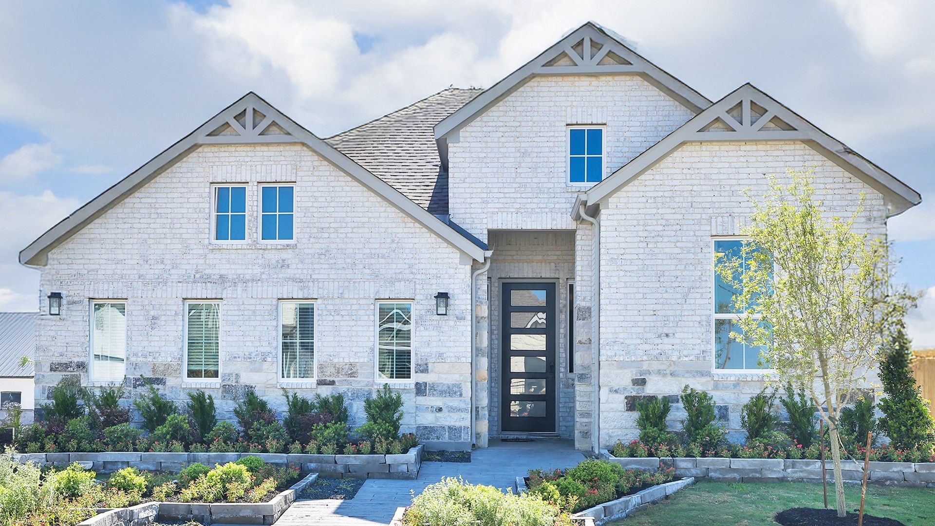 Exquisite white brick facade with gabled roof and geometric door, surrounded by lush landscaping in Southern Pointe.