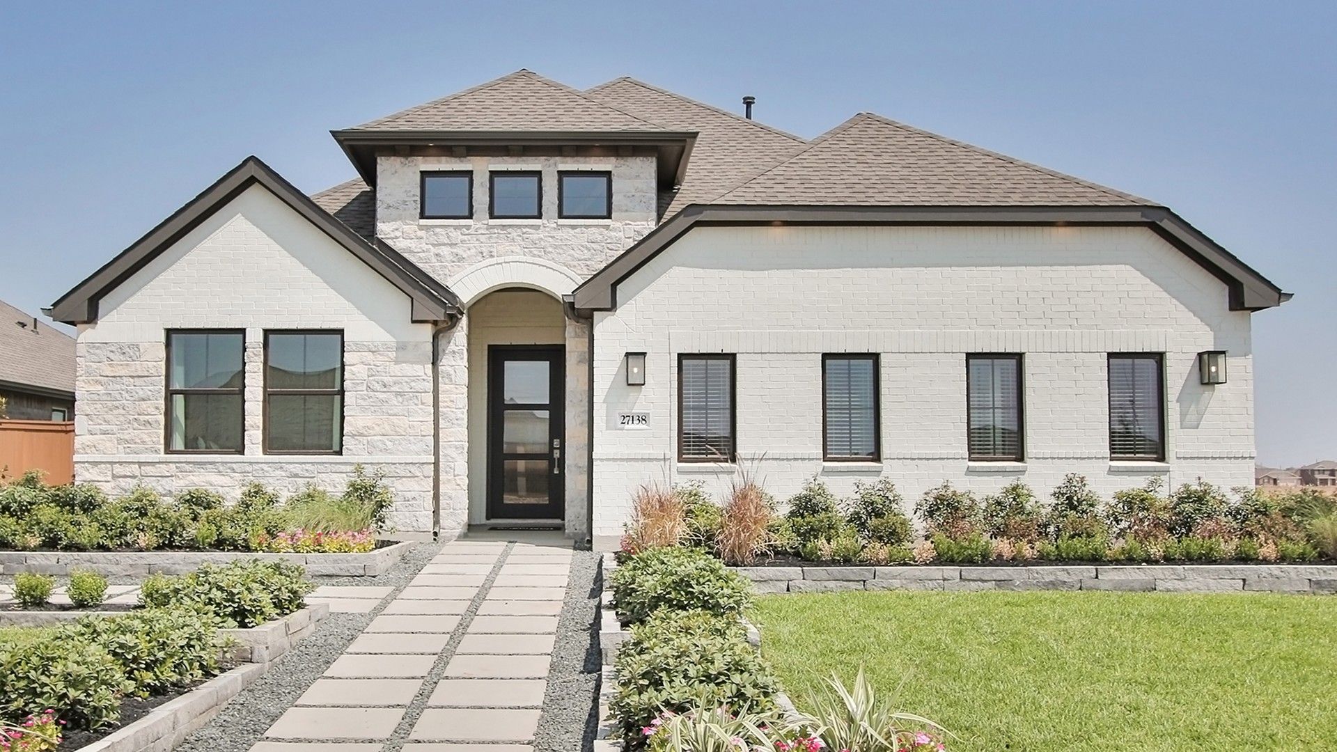 Experience elegance with this Sunterra home, featuring a pristine white brick facade and lush, manicured landscaping.