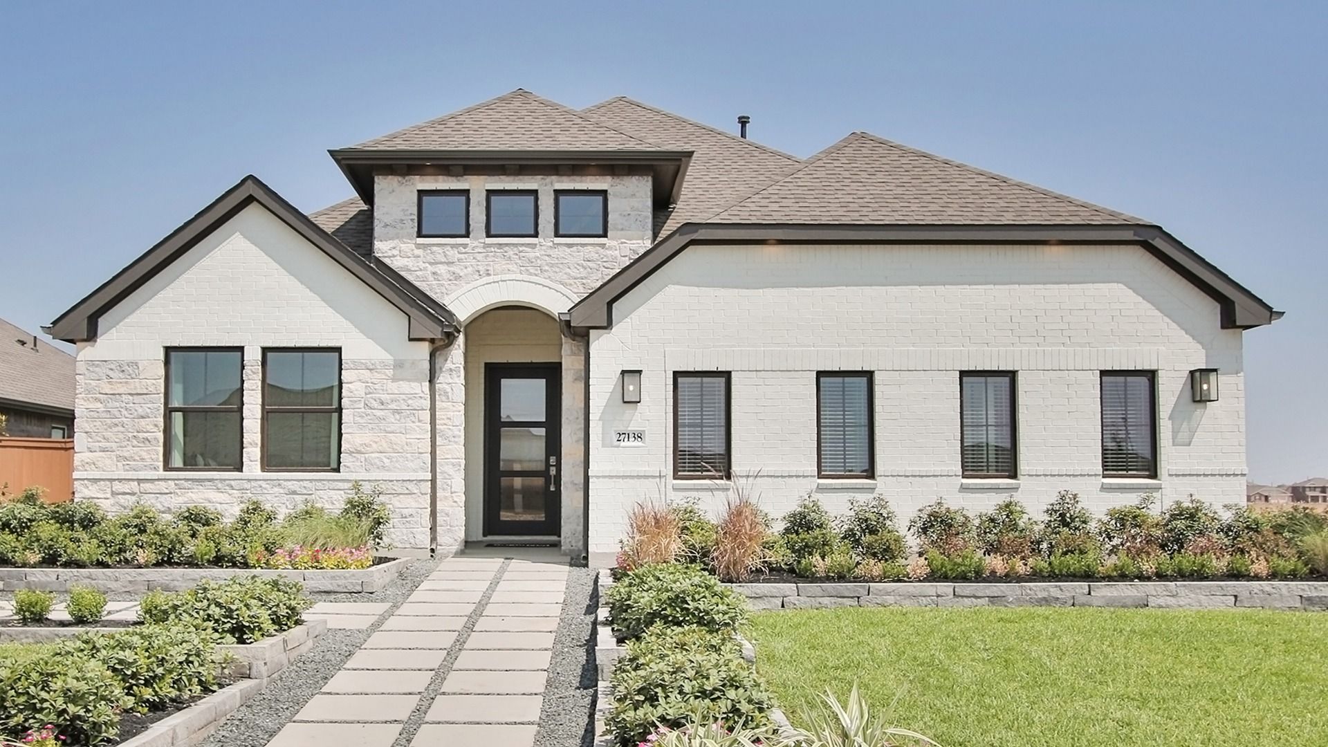 Experience elegance with this Sunterra home, featuring a pristine white brick facade and lush, manicured landscaping.
