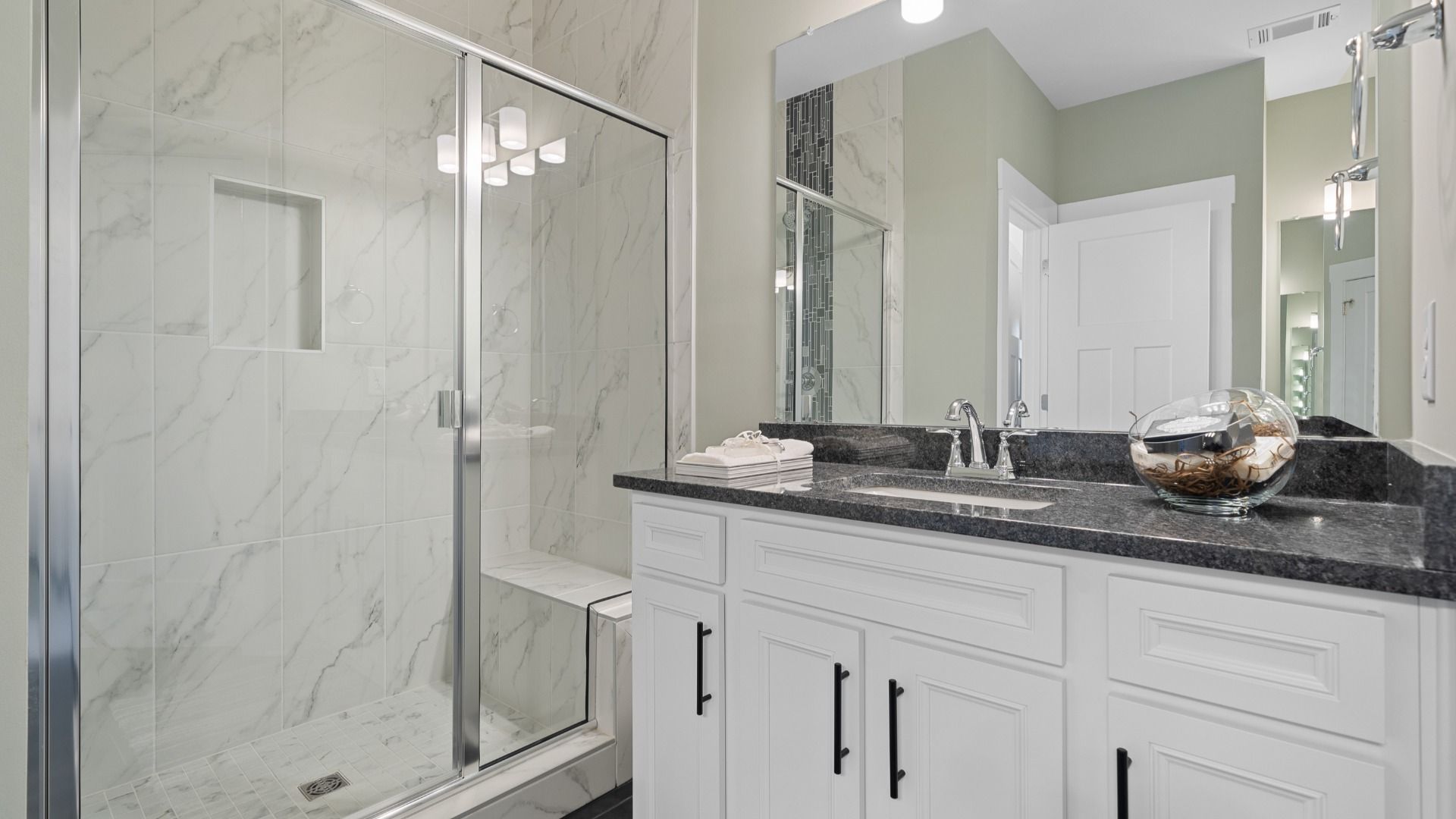 Primary suite bathroom with bright tile shower, dark countertop, white cabinets with dark accent and LVP at Village at Midway