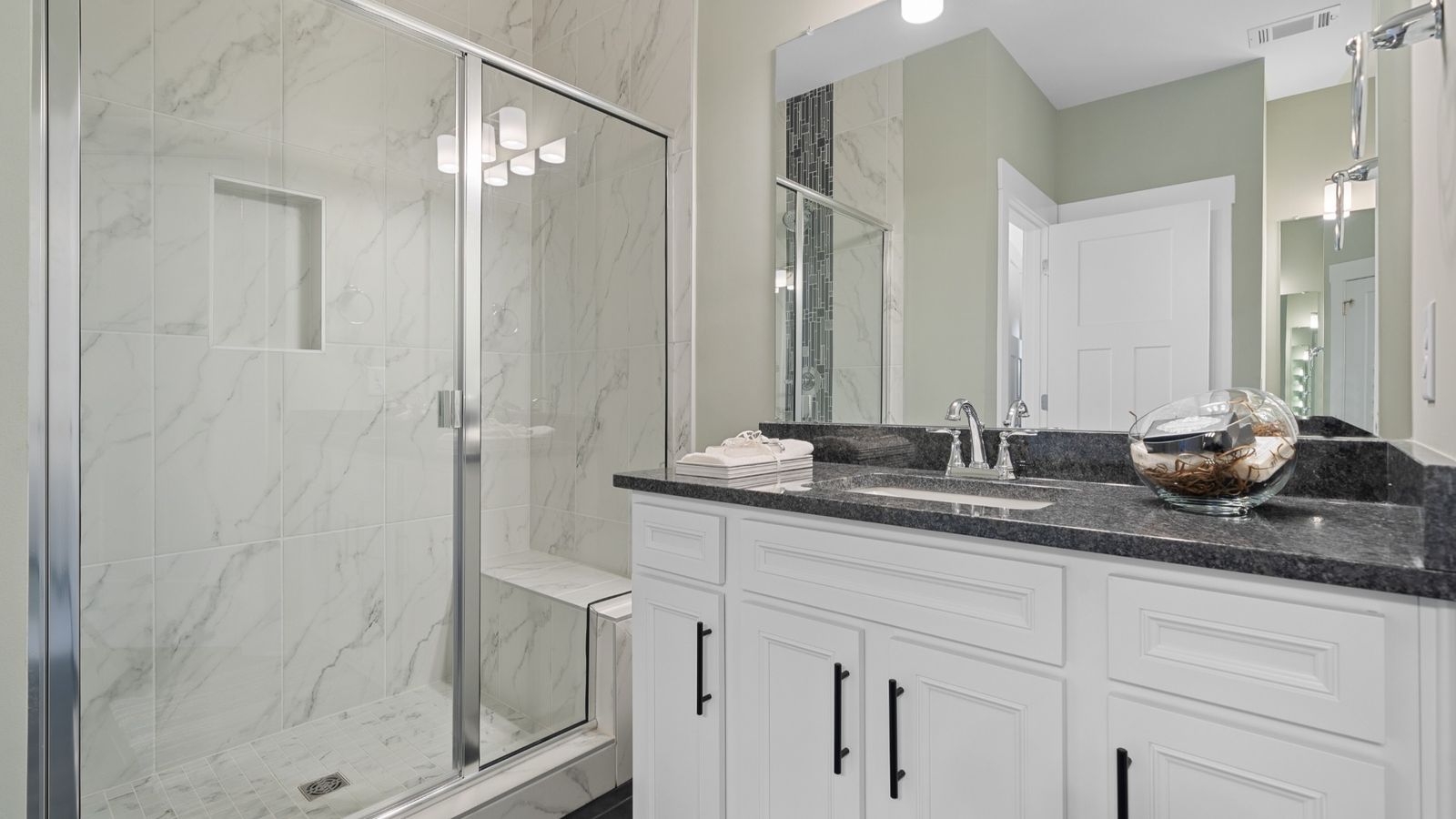 Primary suite bathroom with bright tile shower, dark countertop, white cabinets with dark accent and LVP at Village at Midway