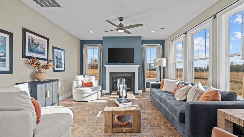 Gorgeous window adorned family room with blue accent wall with fireplace and ceiling fan at Pelham Crossing by DRB Homes