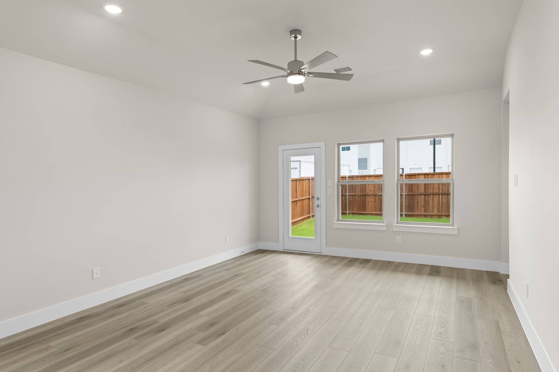 Spacious interior room with wood flooring and ceiling fan, overlooking fenced backyard.