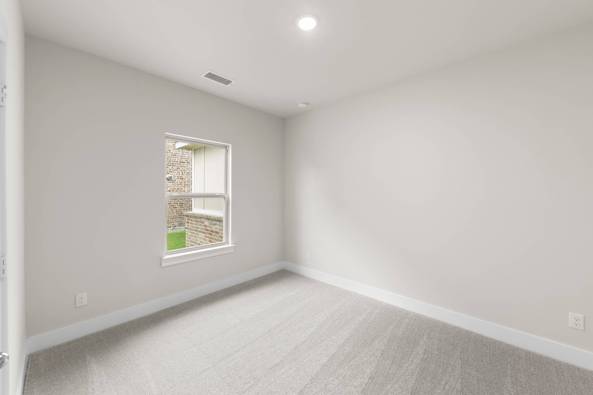 Bright, empty bedroom with neutral walls and carpet, single window view of brick wall.