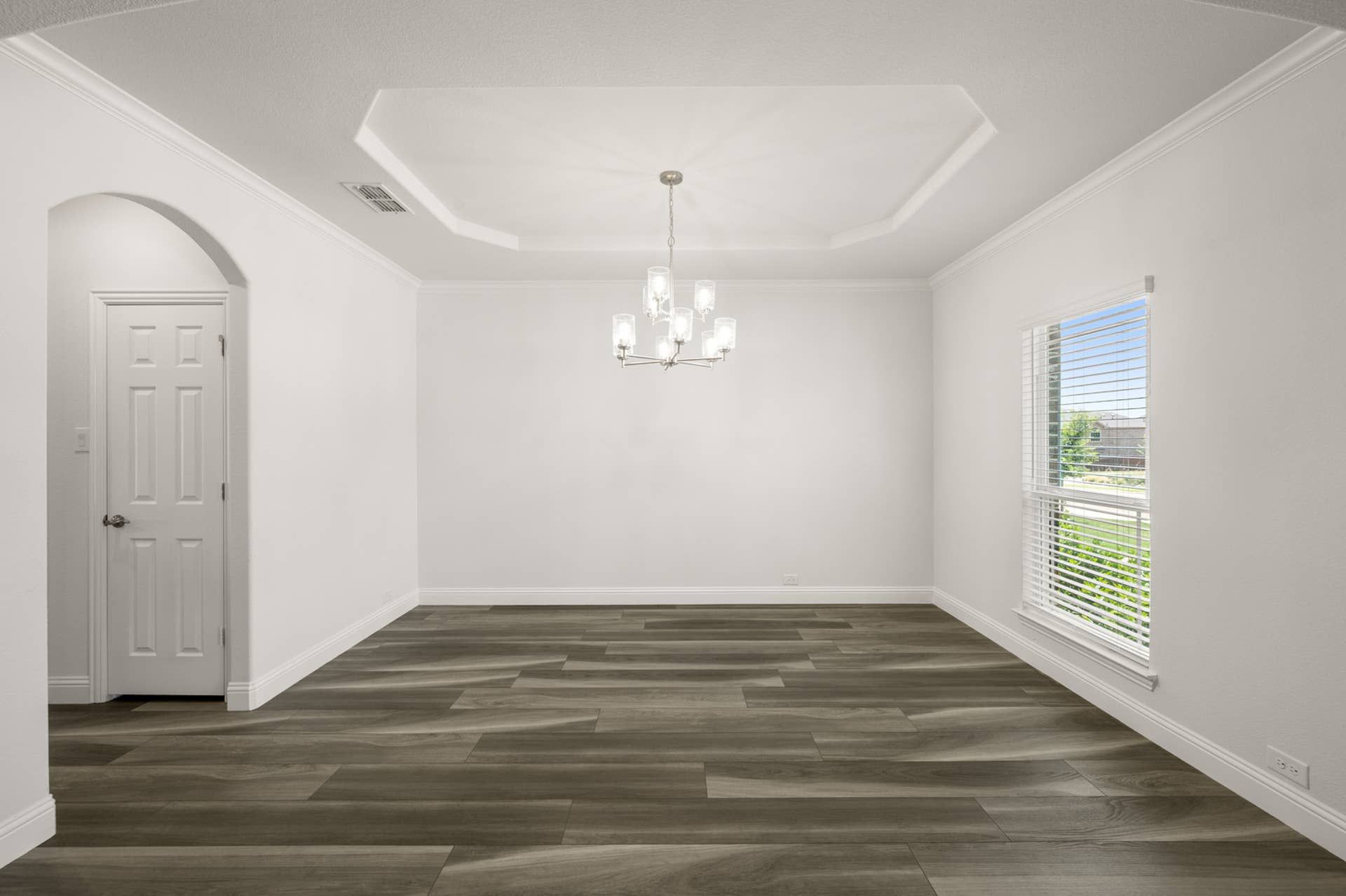 Modern dining room with tray ceiling and elegant chandelier, featuring large window and hardwood floors.