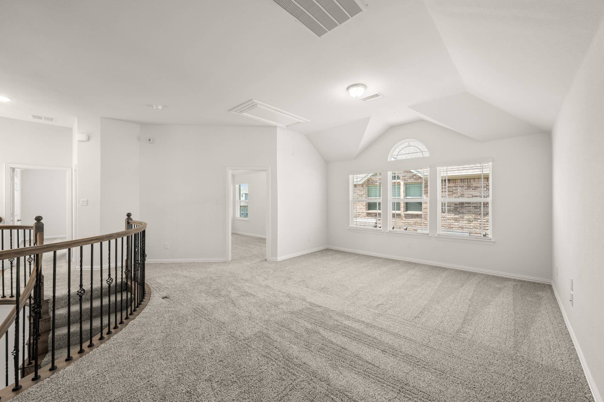 Spacious carpeted game room with arched window and wrought iron staircase.