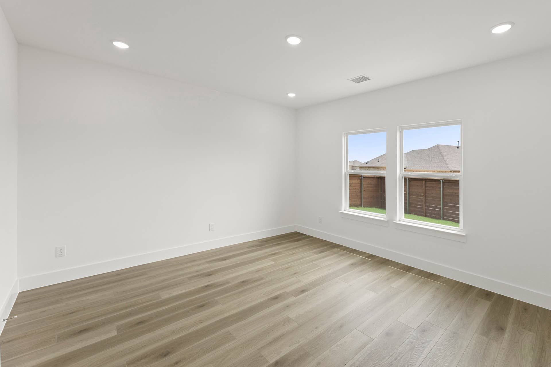 Bright empty room with wood flooring and large windows overlooking a fenced yard.