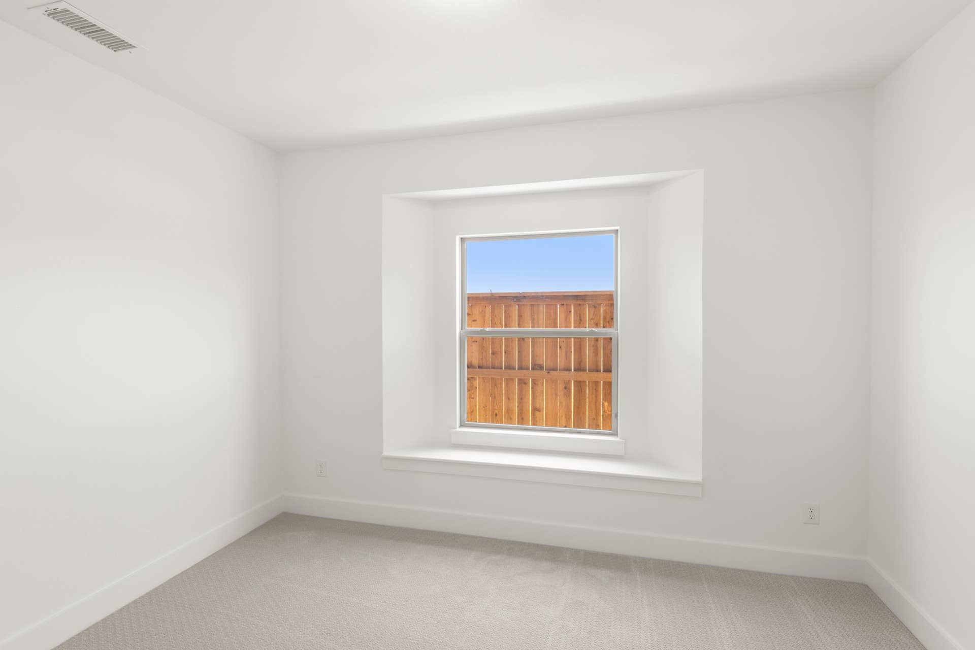 Bright, minimalistic room with carpet and large window facing wooden fence.
