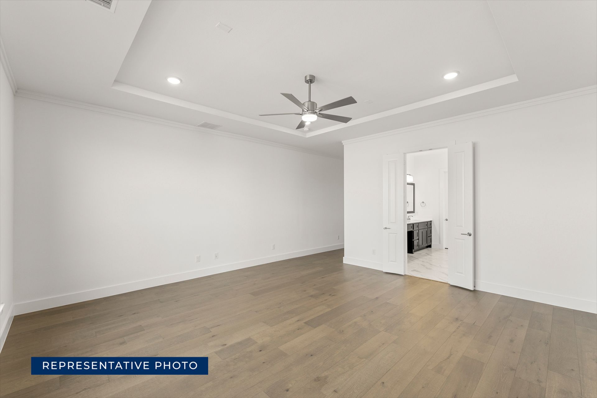 Spacious empty room with wood flooring and ceiling fan, leading to a bathroom.