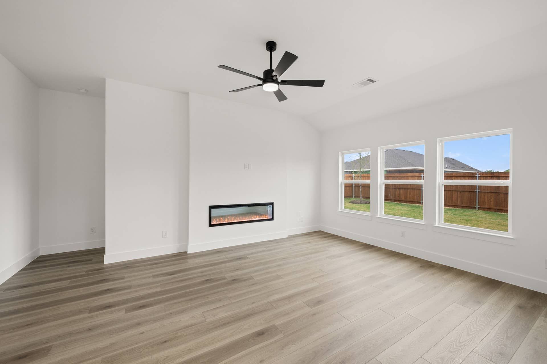 Modern living room with fireplace, ceiling fan, and large windows overlooking backyard.