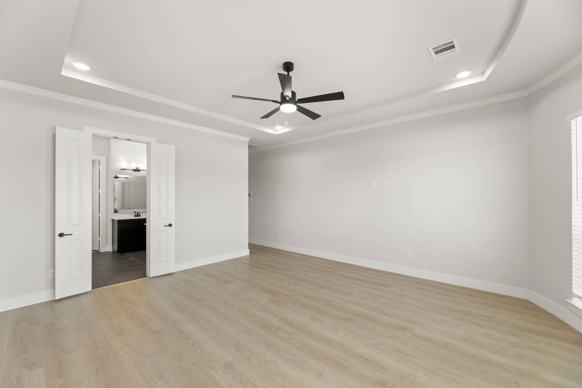Spacious modern interior with wood floors, ceiling fan, and double doors to bathroom.