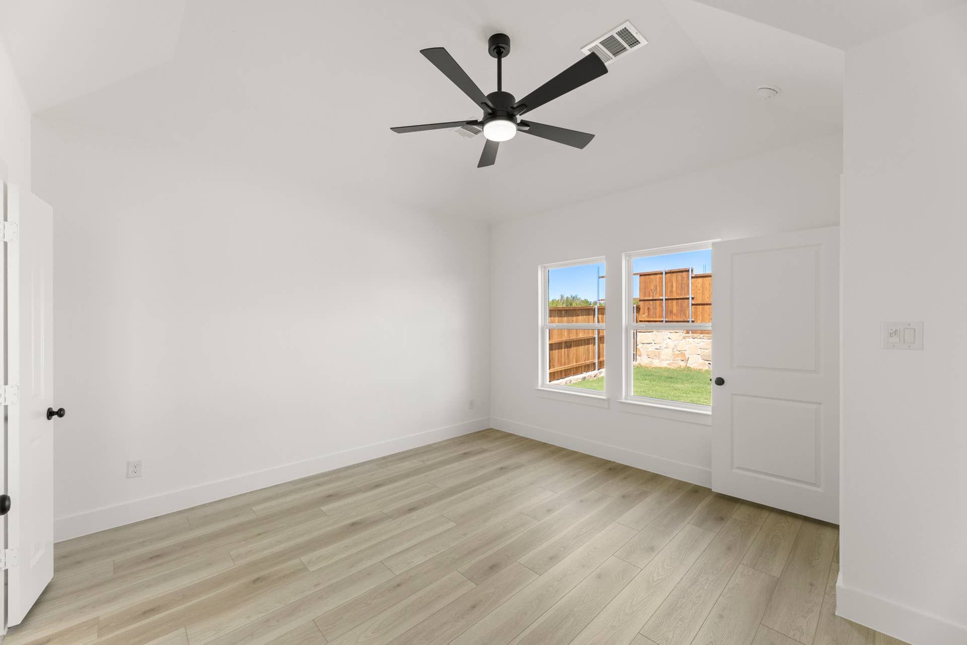 Bright room with light wood flooring and ceiling fan, overlooking fenced backyard.