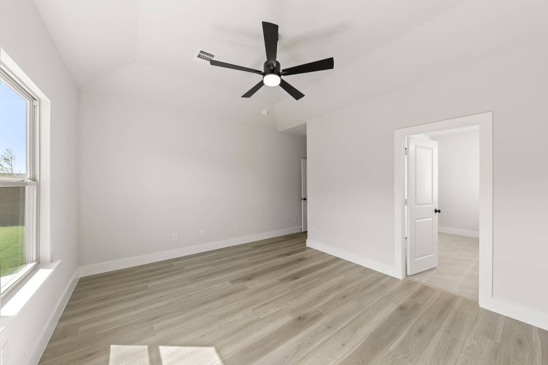 Spacious room with light wood flooring and ceiling fan, adjacent to a bright doorway.