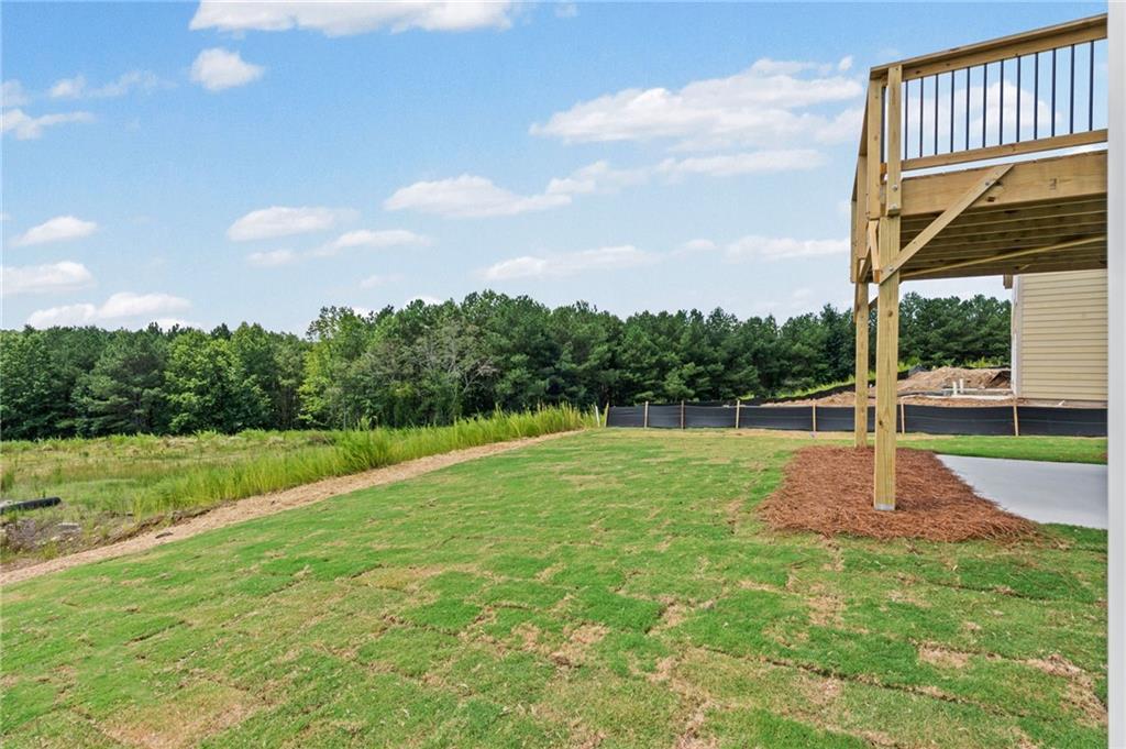 Expansive green space featuring a wooden deck with black balusters, a concrete patio, and a natural treeline backdrop