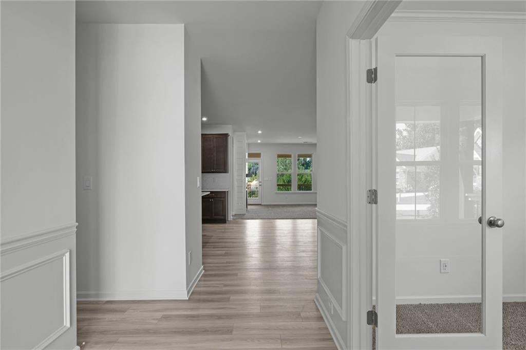 Bright interior hallway featuring wood-finish flooring, white paneled wainscoting, a glass-paneled door, recessed lighting, and a kitchen area with dark wood cabinetry