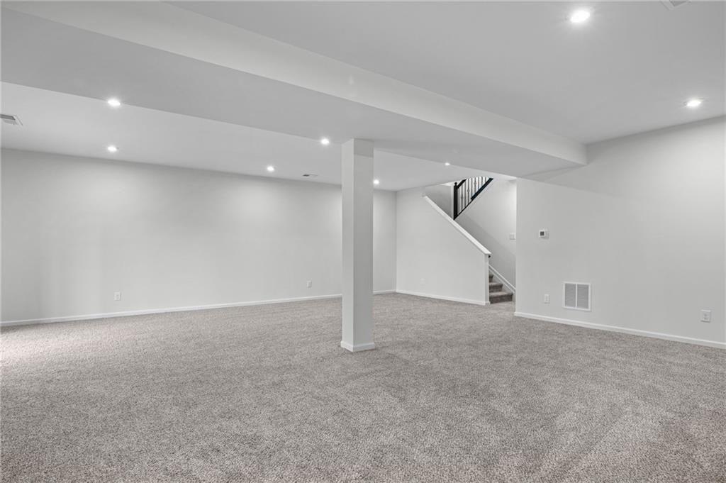 Spacious open-concept lower level featuring plush grey carpeting, recessed lighting, white painted walls, a structural support column, and a staircase with black metal balusters