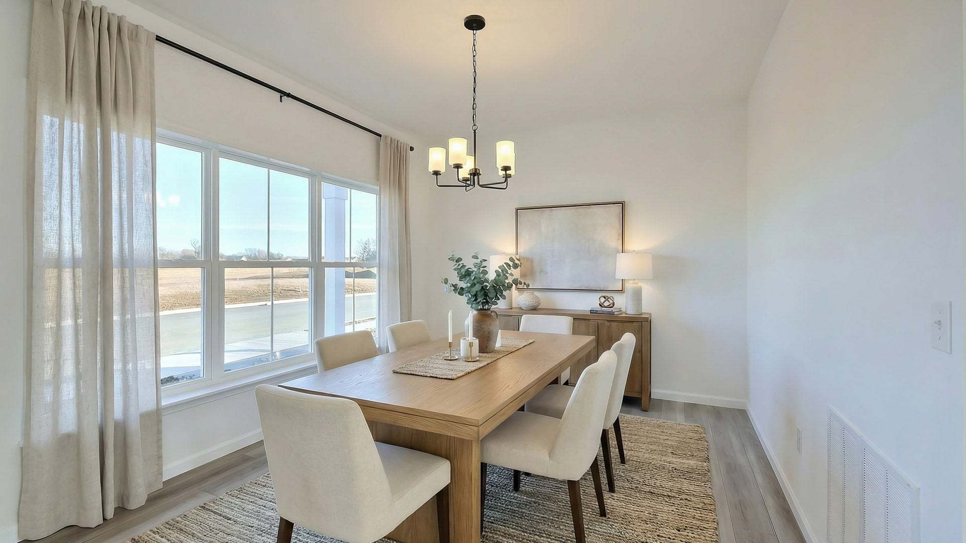 Modern dining room with wooden table, white chairs, and chandelier near large windows.