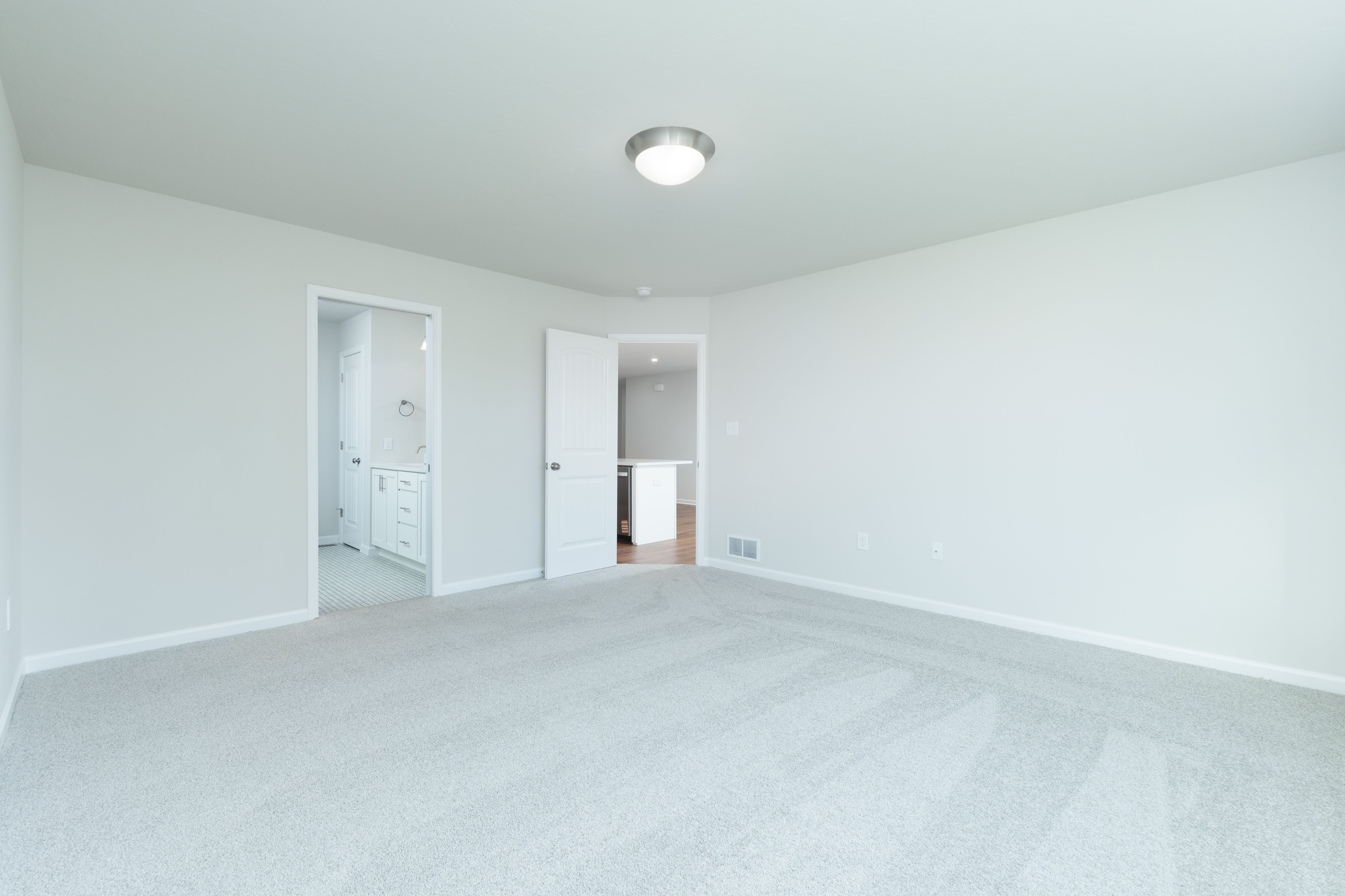 Spacious owners bedroom with light gray walls, plush carpet, and open door to bath