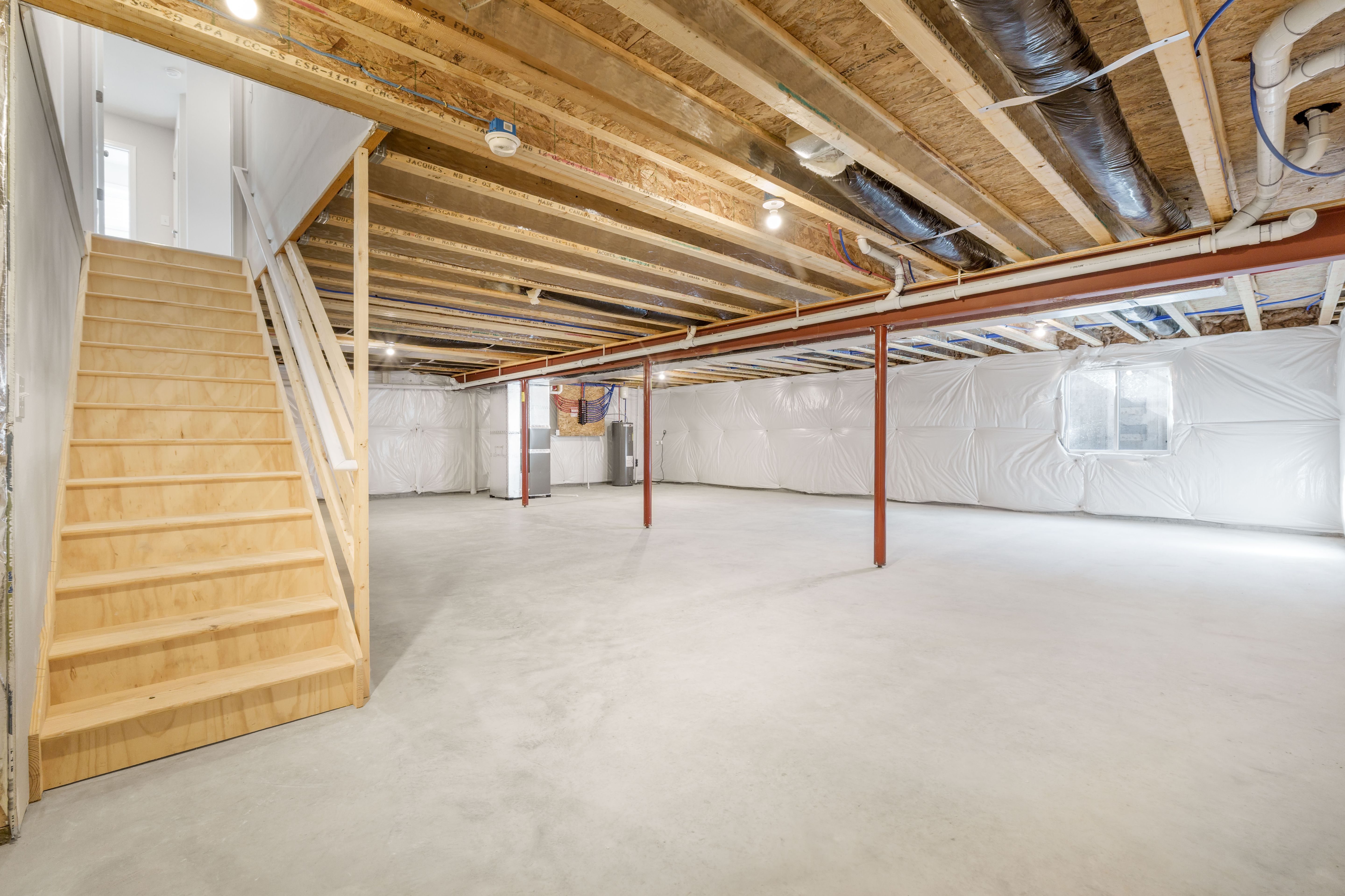 Unfinished basement with concrete floor, insulated walls, exposed ceiling beams, and wood staircase