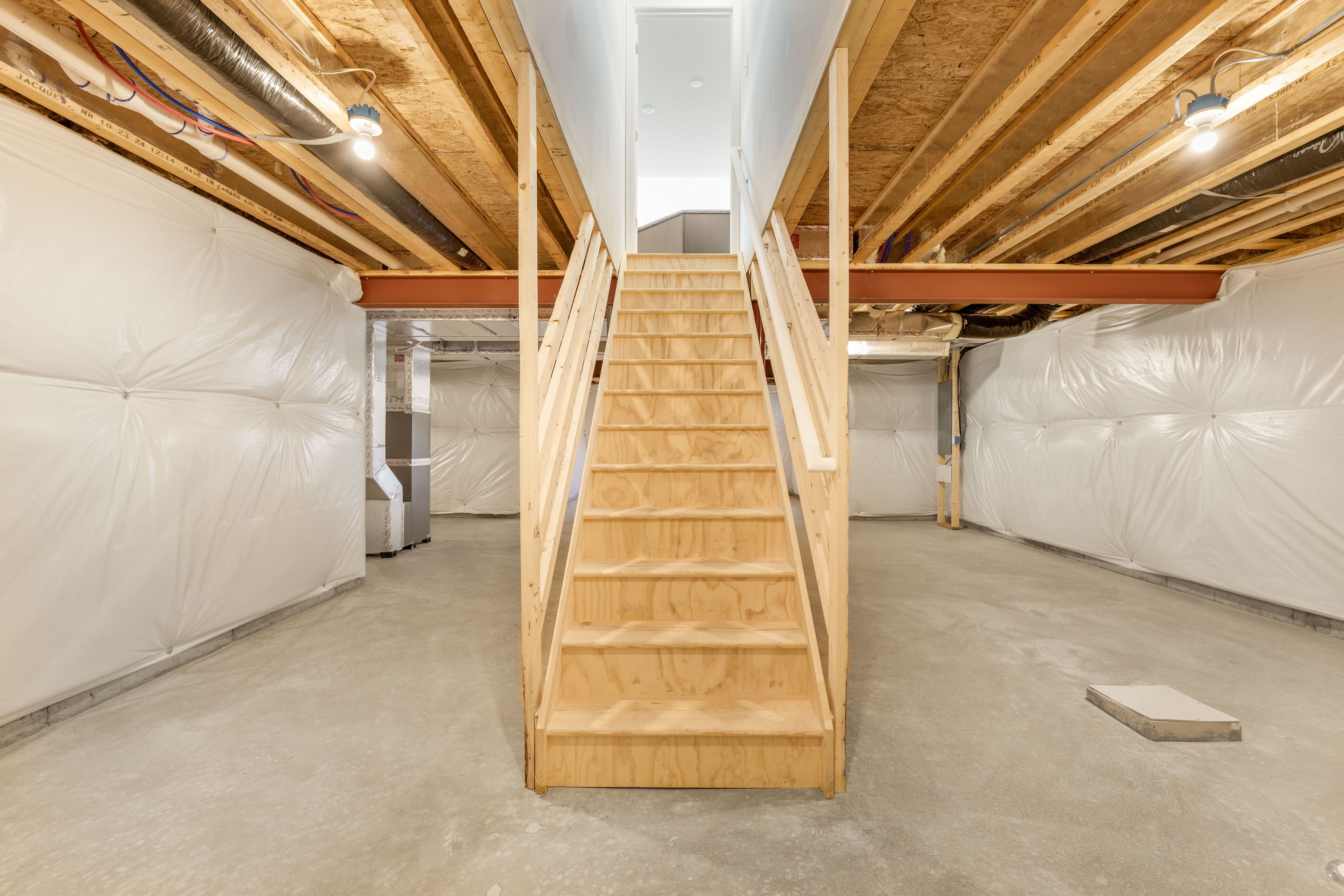 Unfinished basement with wood staircase, concrete floors, insulated walls, and exposed ceiling beams with lighting.