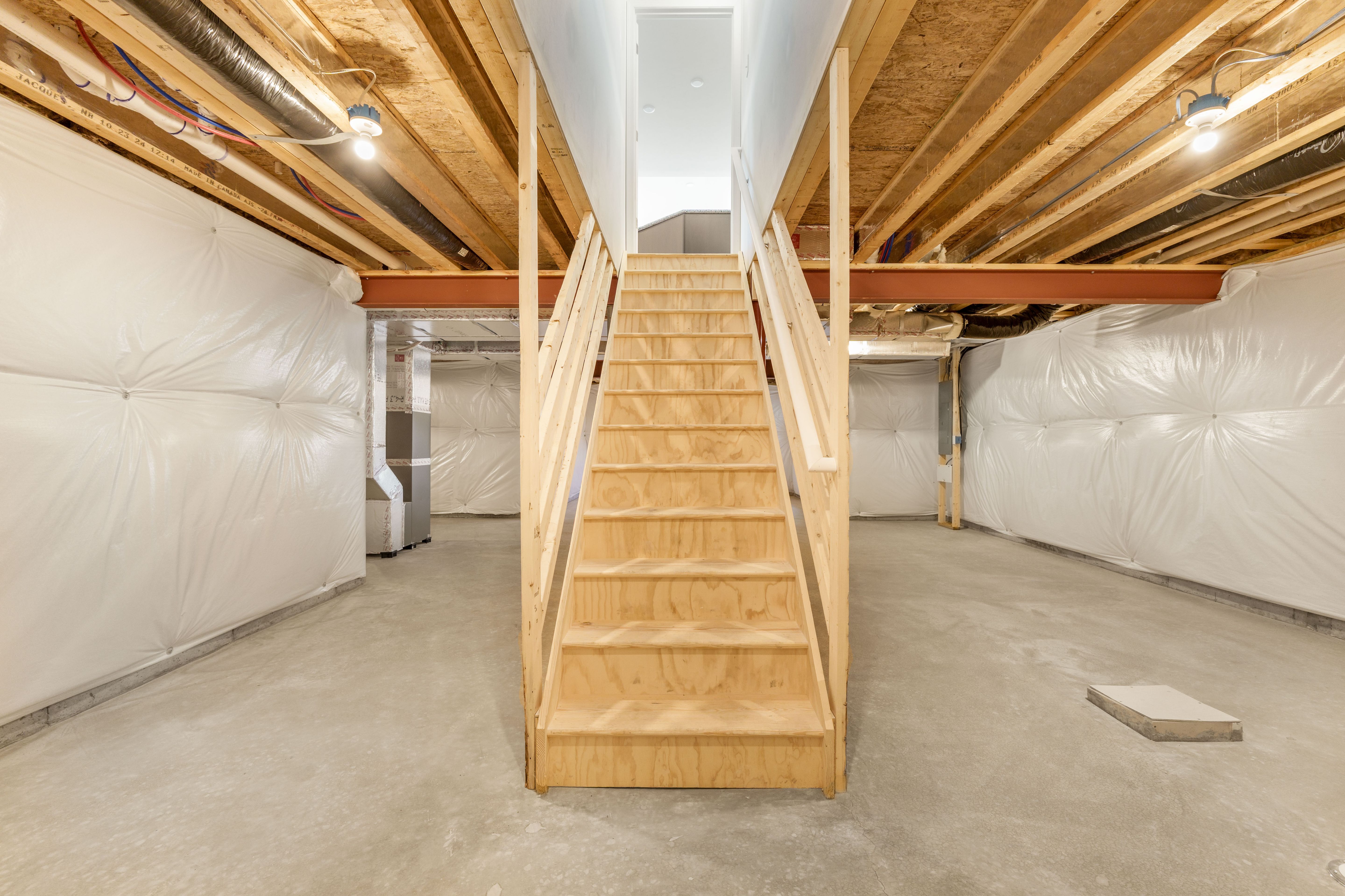 Unfinished basement with wood staircase, concrete floors, insulated walls, and exposed ceiling beams with lighting.