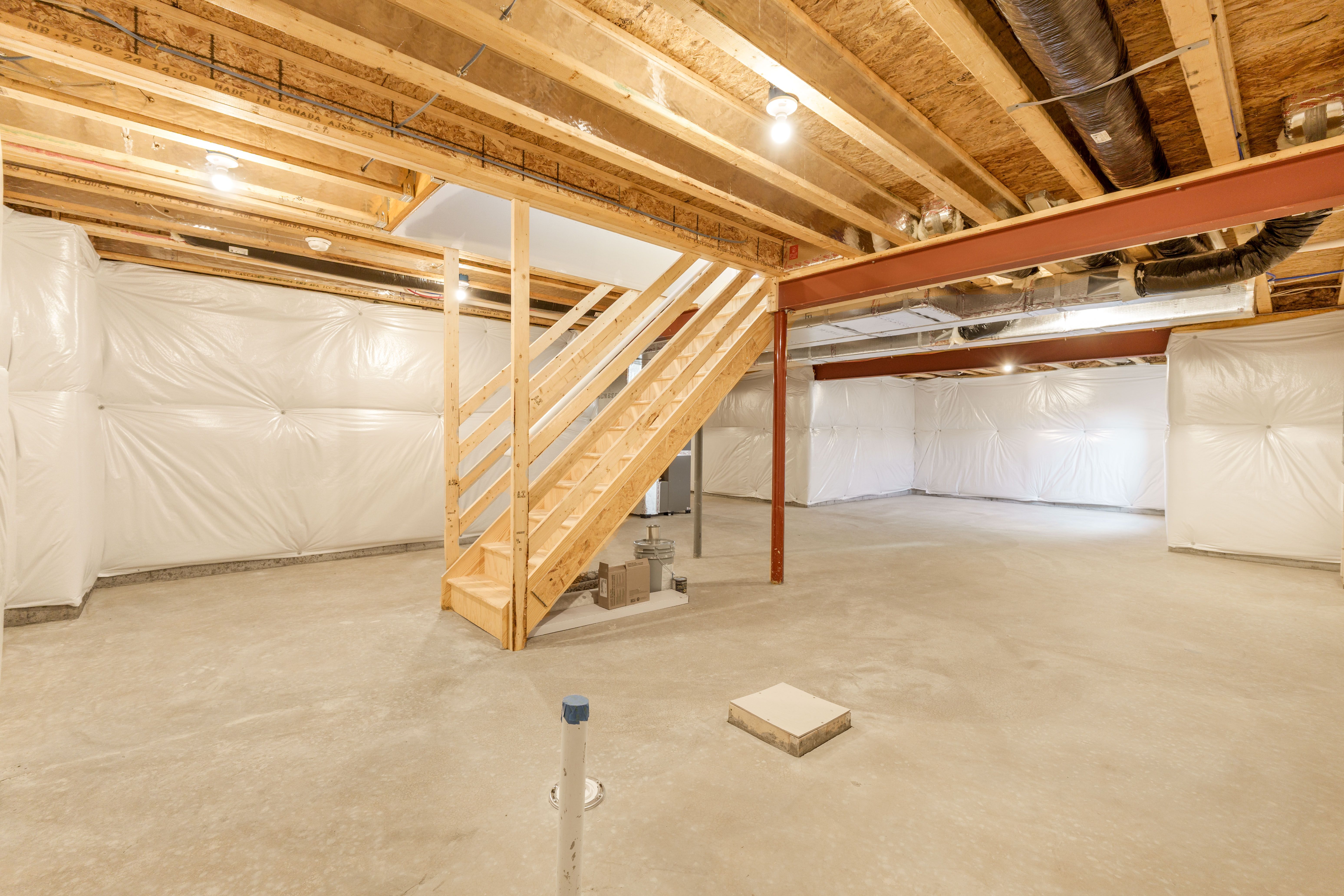 Wide view of unfinished basement with wood stairs, steel support beams, concrete floors, and insulated foundation walls.