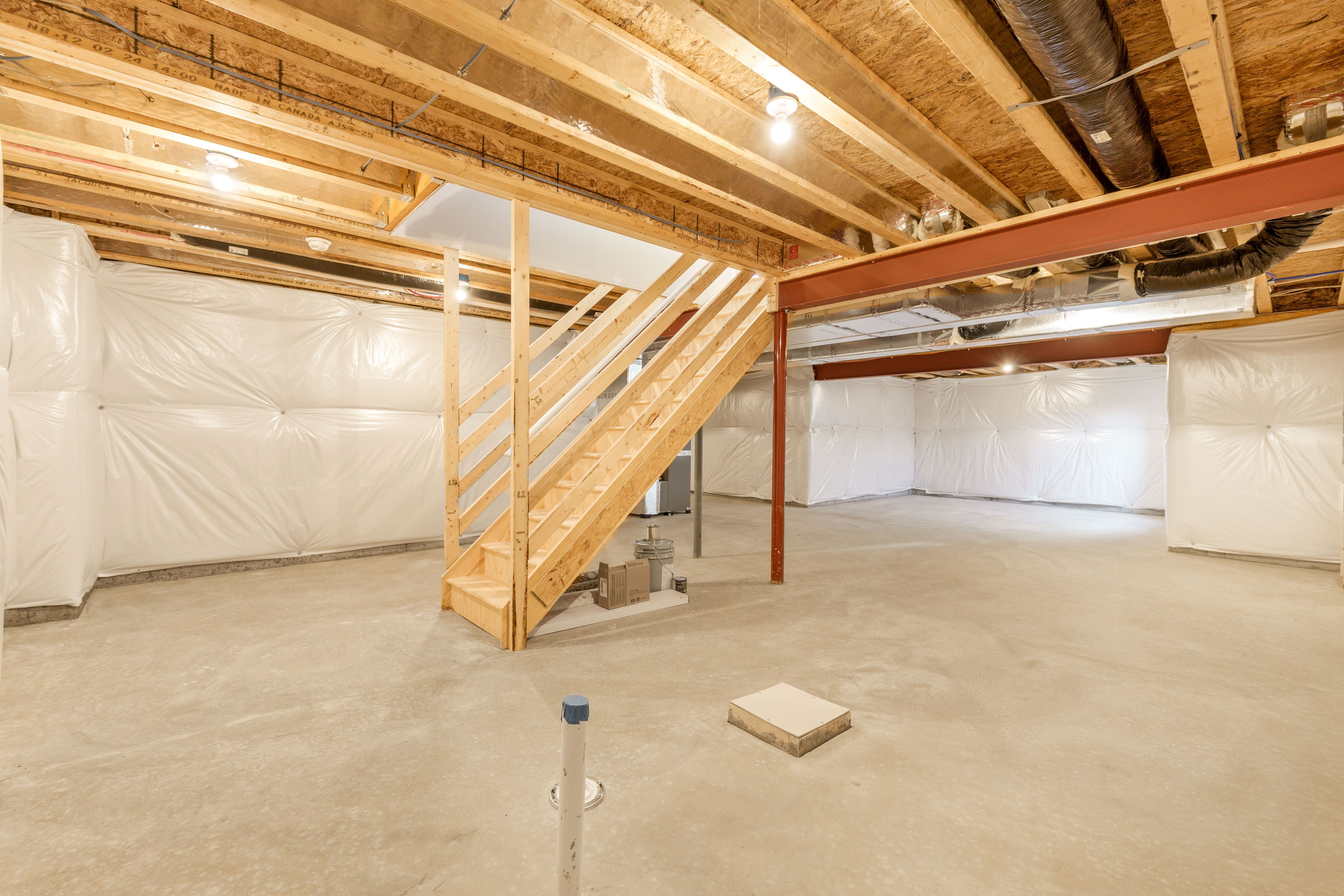 Wide view of unfinished basement with wood stairs, steel support beams, concrete floors, and insulated foundation walls.