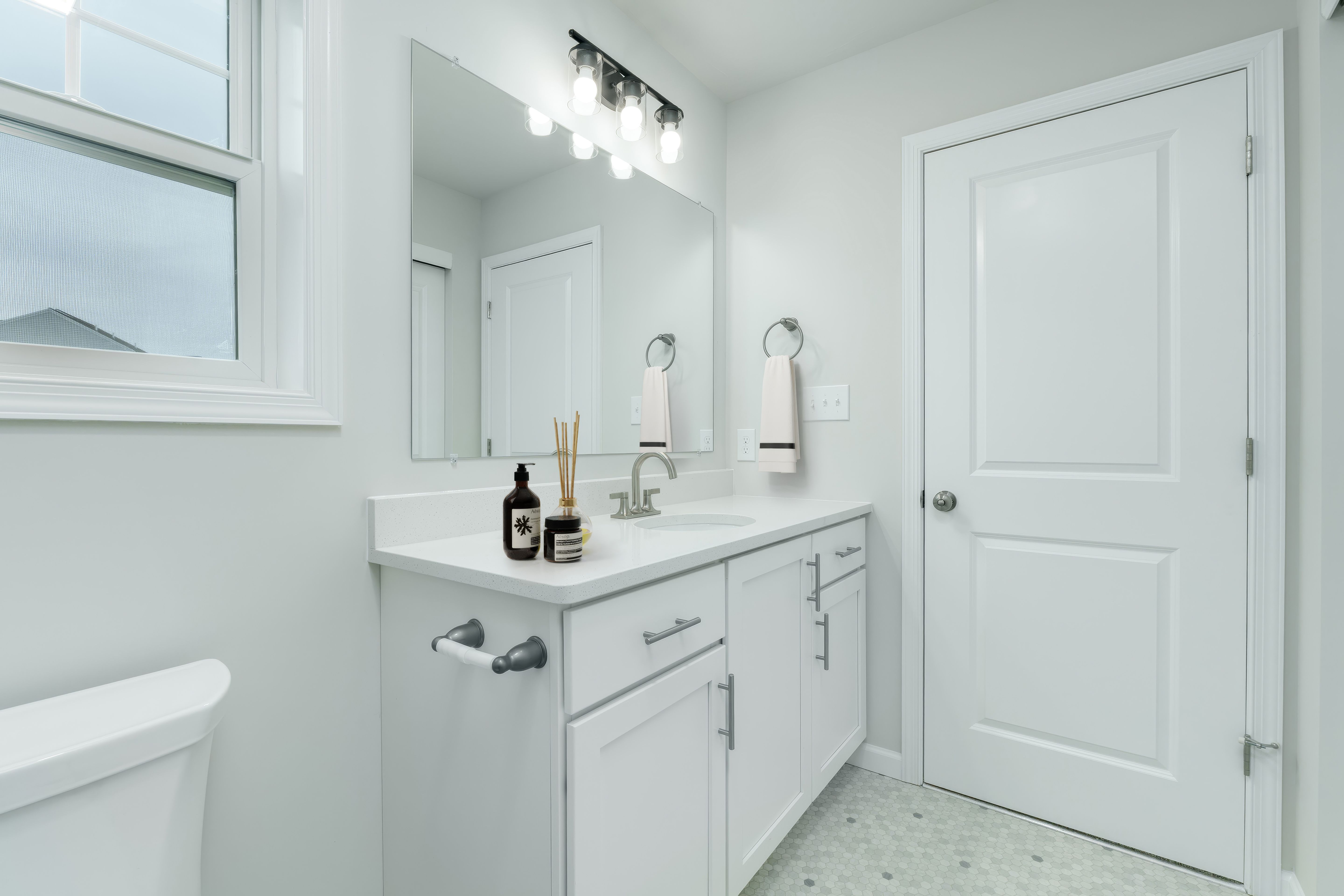 Owners bathroom with white vanity, large mirror, modern lighting, and light hex tile flooring.