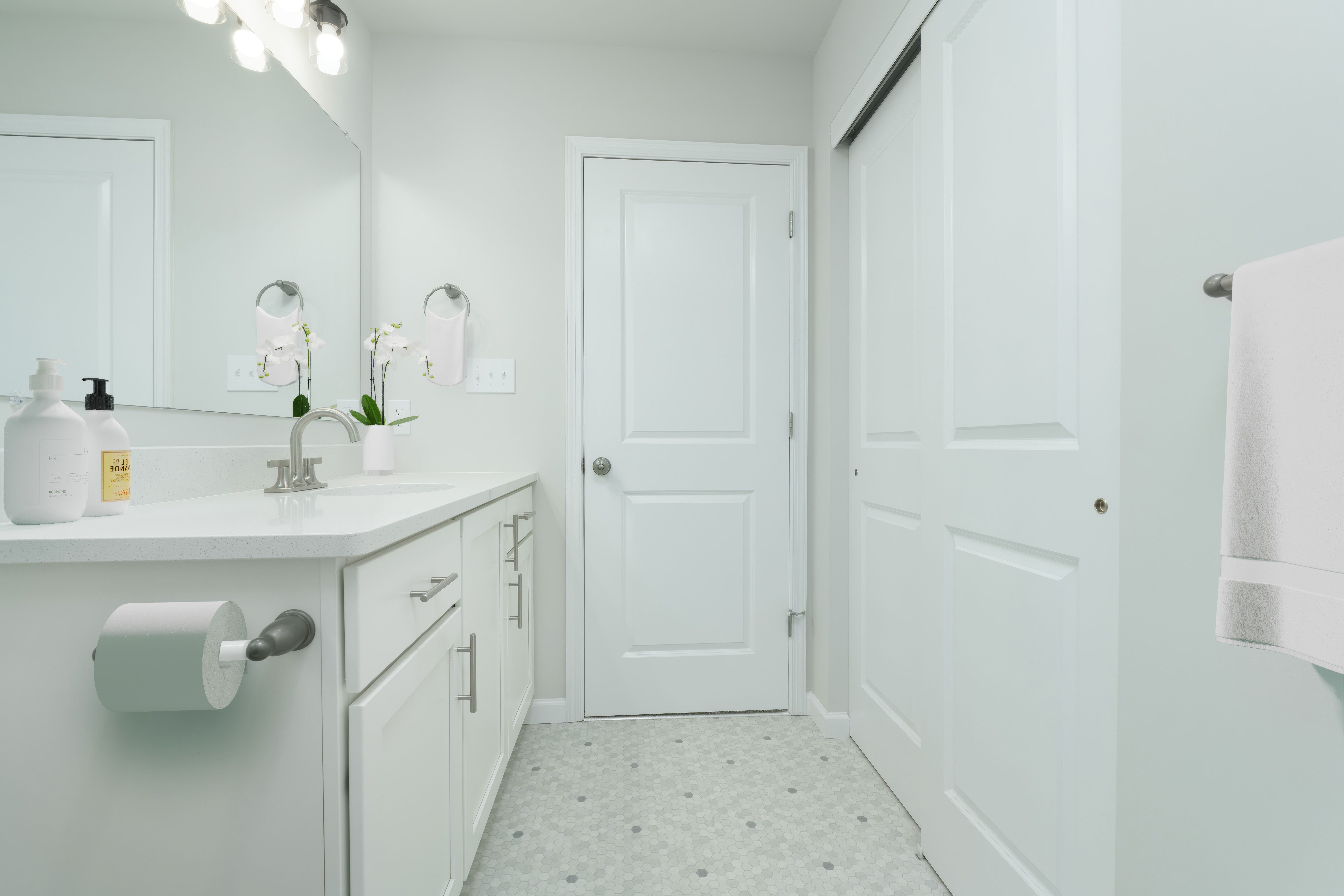 Bright bathroom vanity with white cabinets, wide mirror, modern lighting, and door to closet.