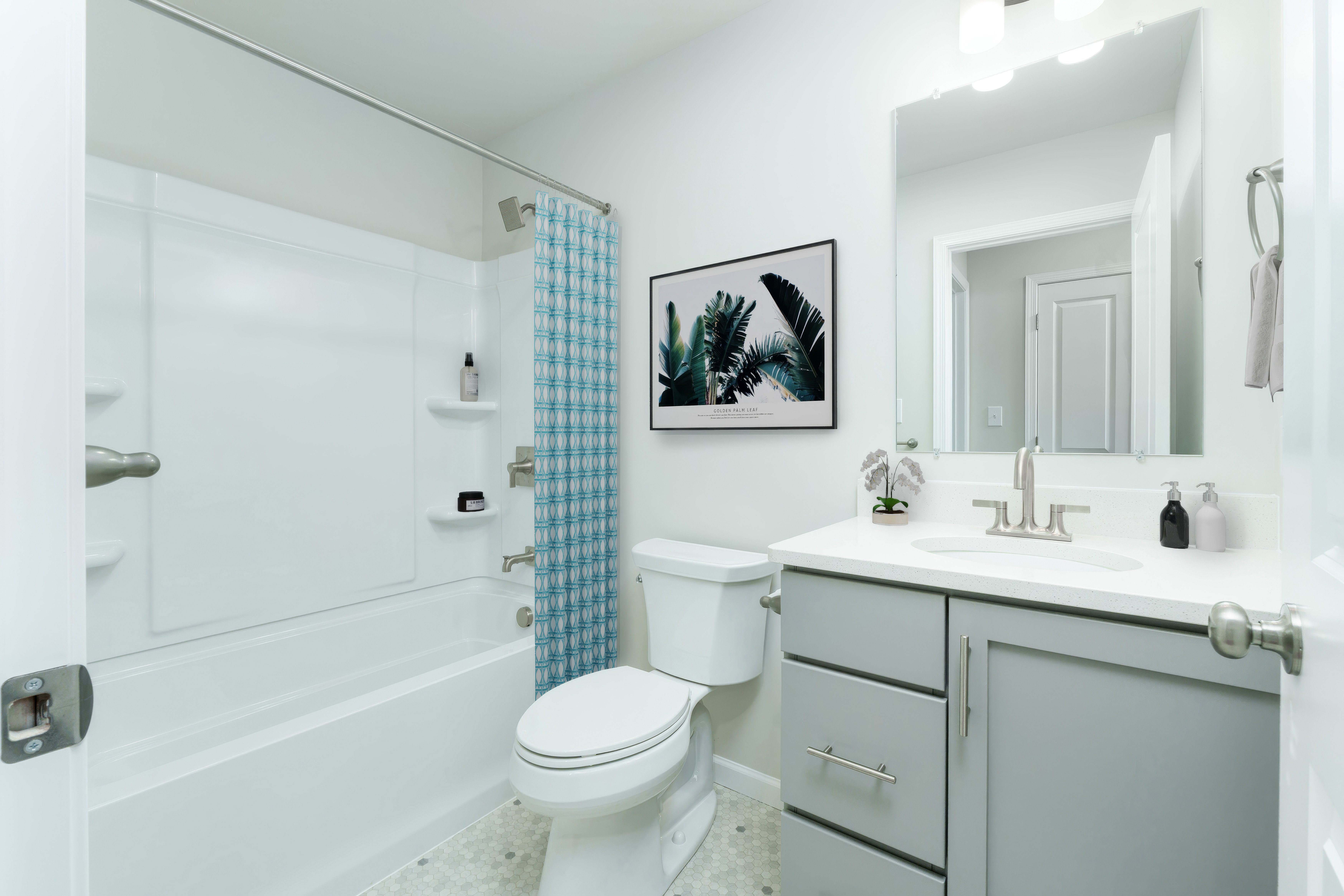 Bright bathroom with white tub and shower surround, patterned blue curtain, toilet, and gray vanity with sink and large mirror.