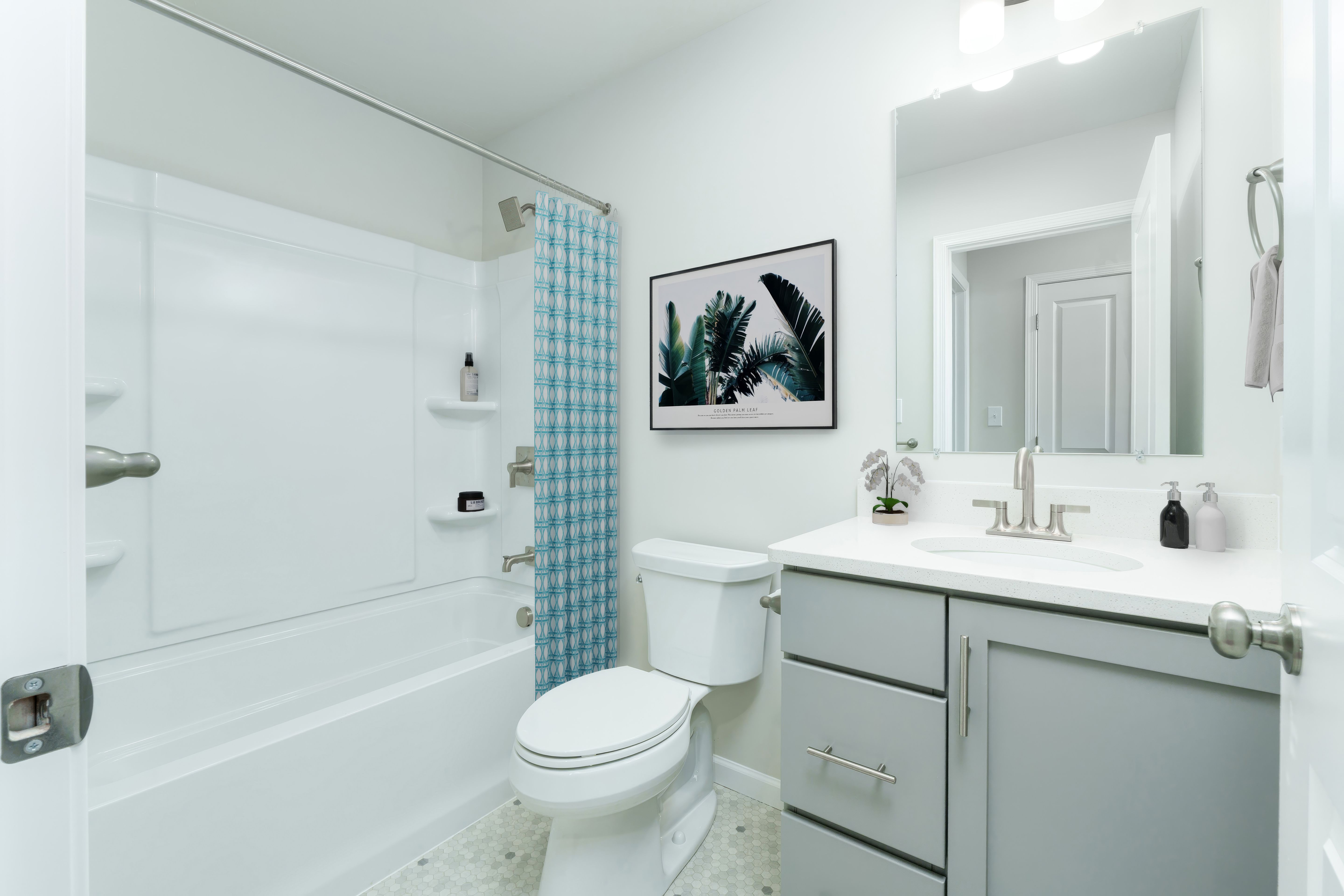 Bright bathroom with white tub and shower surround, patterned blue curtain, toilet, and gray vanity with sink and large mirror.