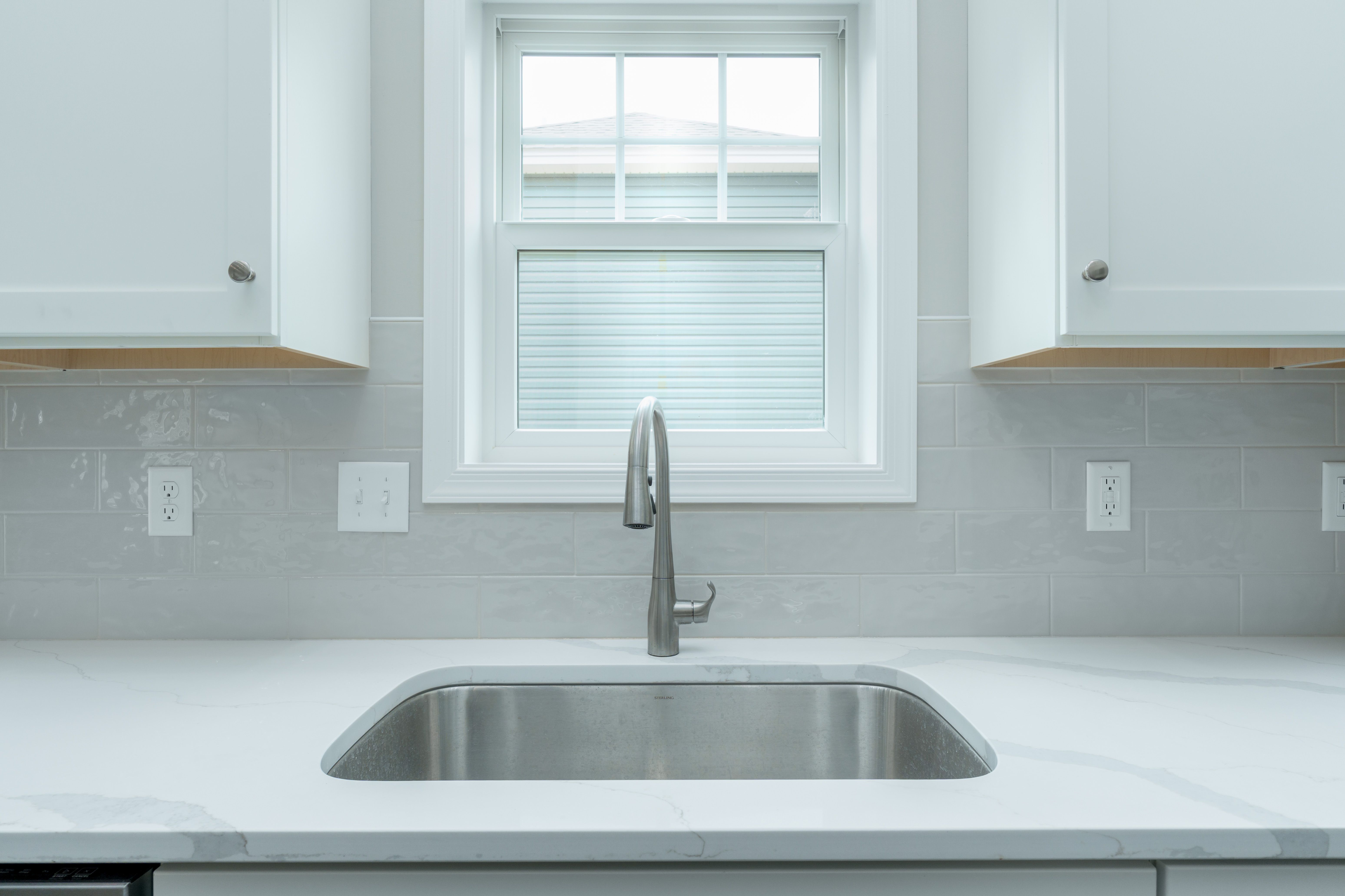 Kitchen sink beneath bright window with white quartz countertops and glossy tile backsplash