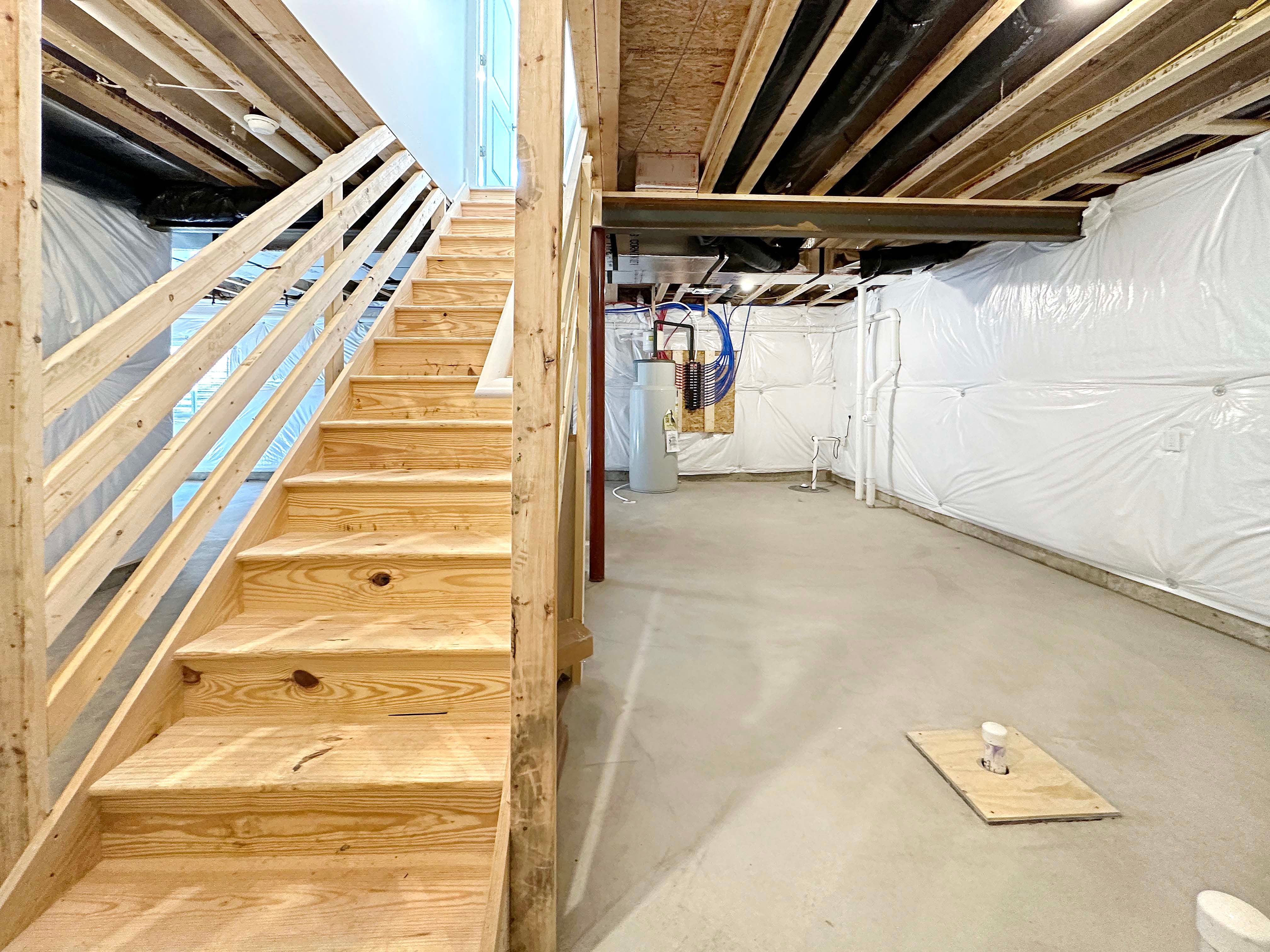 Unfinished basement- wood stairs concrete floor, I-joist floor system above, Manabloc on wall in rear.