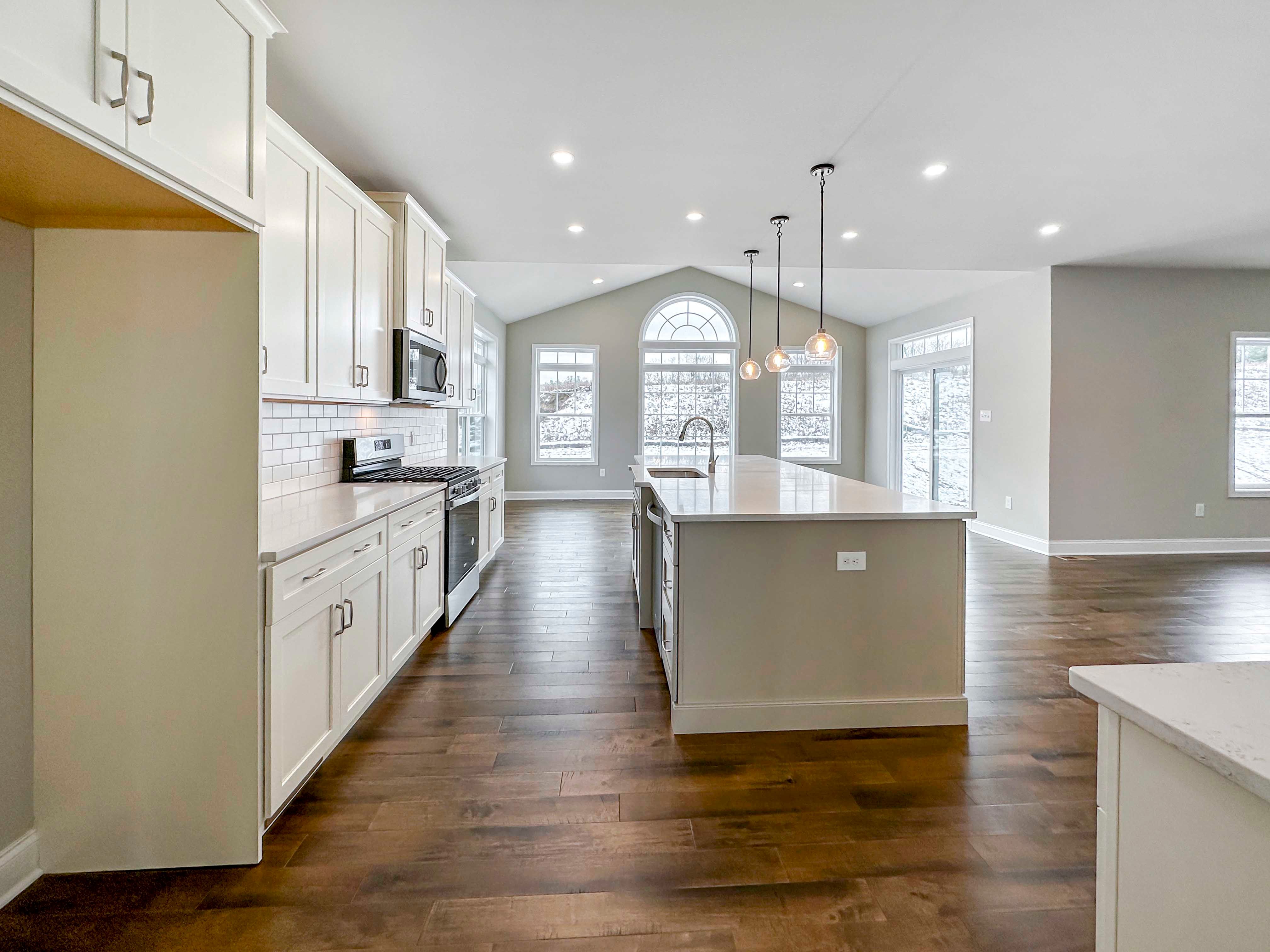 Kitchen - Luxury Kitchen with refrigerator space to left, white cabinetry, white countertops, tile backsplash, island with sink and overhead pendant lighting, and stainless appliances - with the Opt. Keeping Room in the rear.