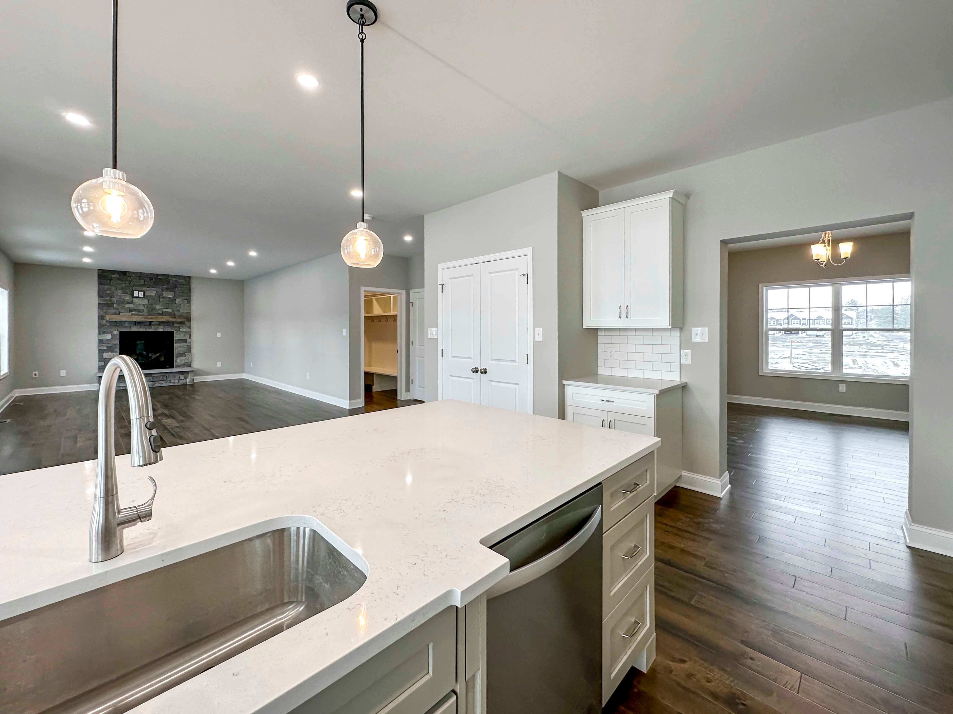 Kitchen Island - with white cabinetry, built-in stainless dishwasher, single-basin sink, and overhead pendant lighting. Drop Zone and Pantry just beyond island. Entrance to Dining Room to right, to left is Family Room.