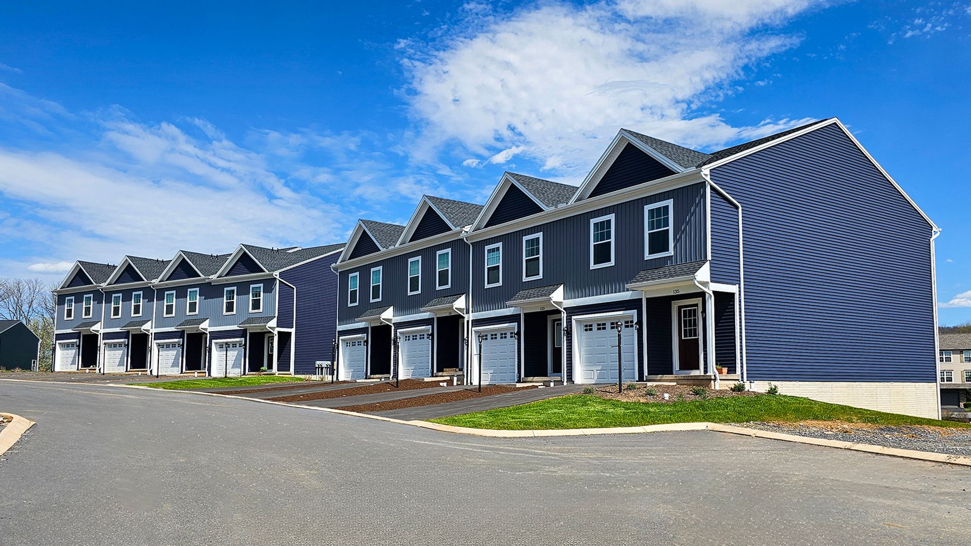 Long row of newly built grey townhouses with white trim, dark red doors, single-car garages, and paved driveways. Bright blue sky.