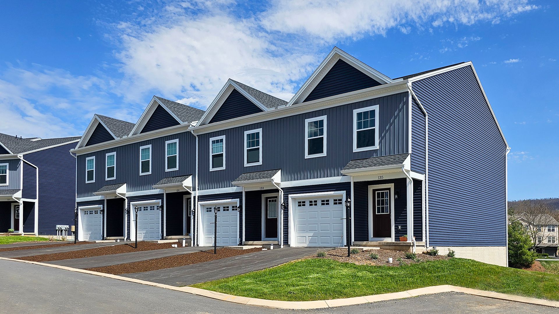 Row of new townhouses with grey siding, white trim, dark red doors, single-car garages, and paved driveways. Clear blue sky