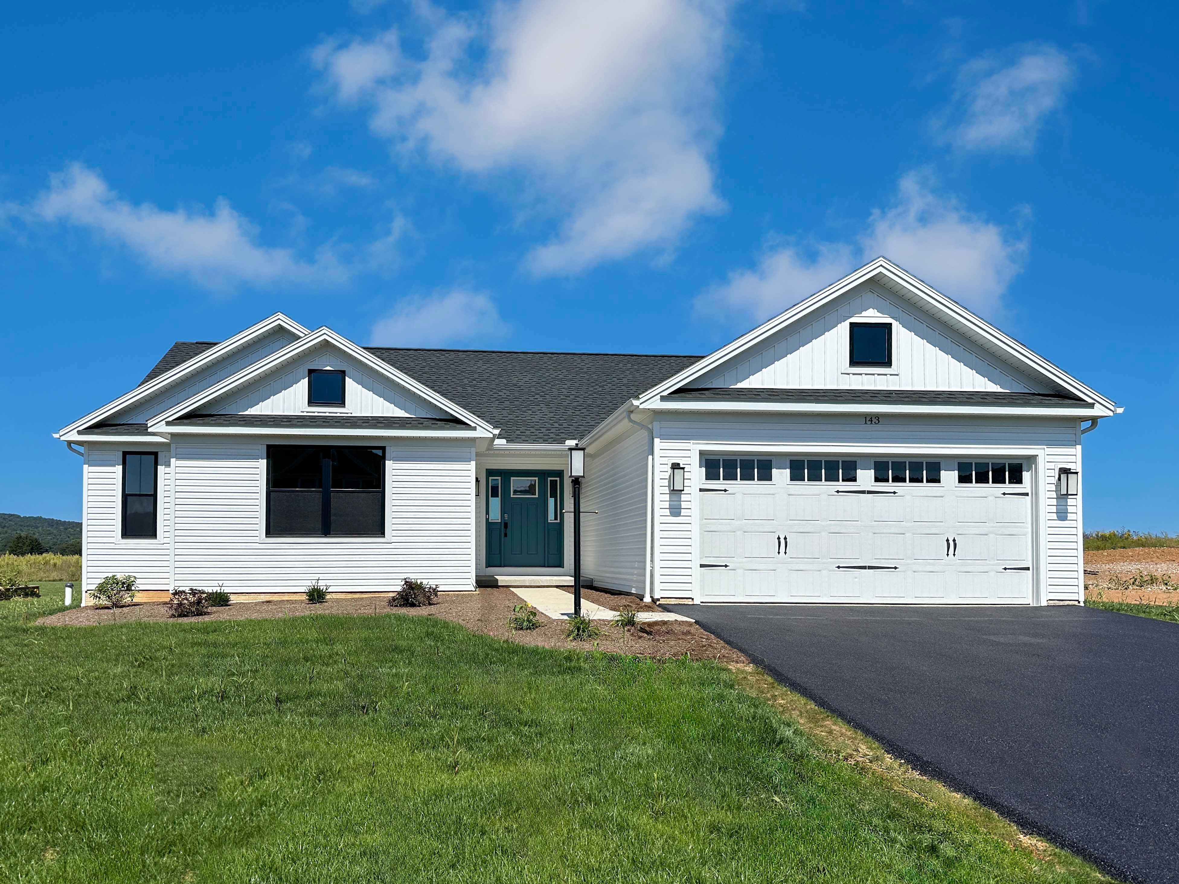White ranch-style home with black roof, teal front door, two-car garage, and landscaped yard, set against a bright blue sky.