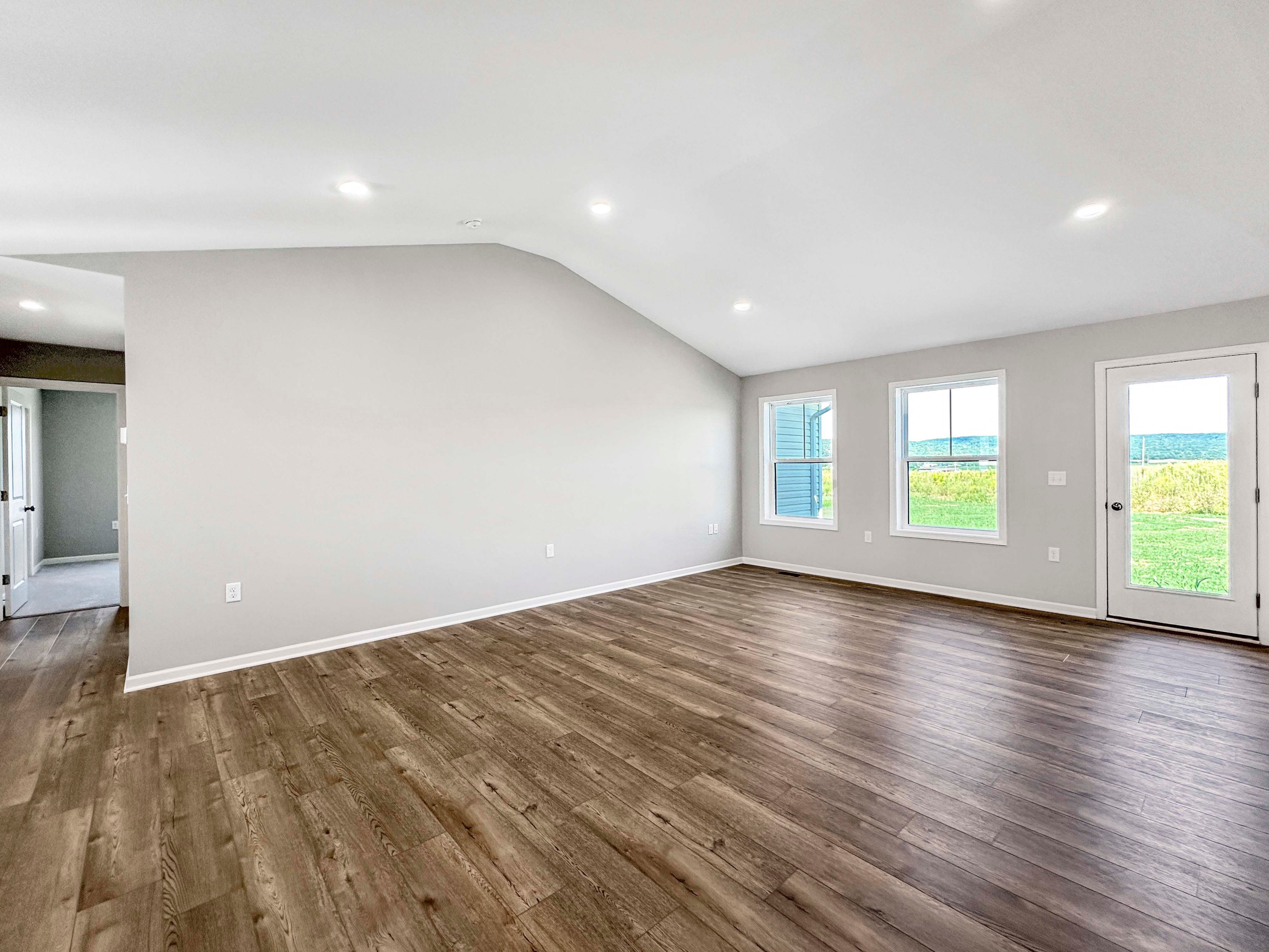 Spacious great room featuring large windows, vaulted ceiling, and a door leading to the outside. Wood-like floors throughout.