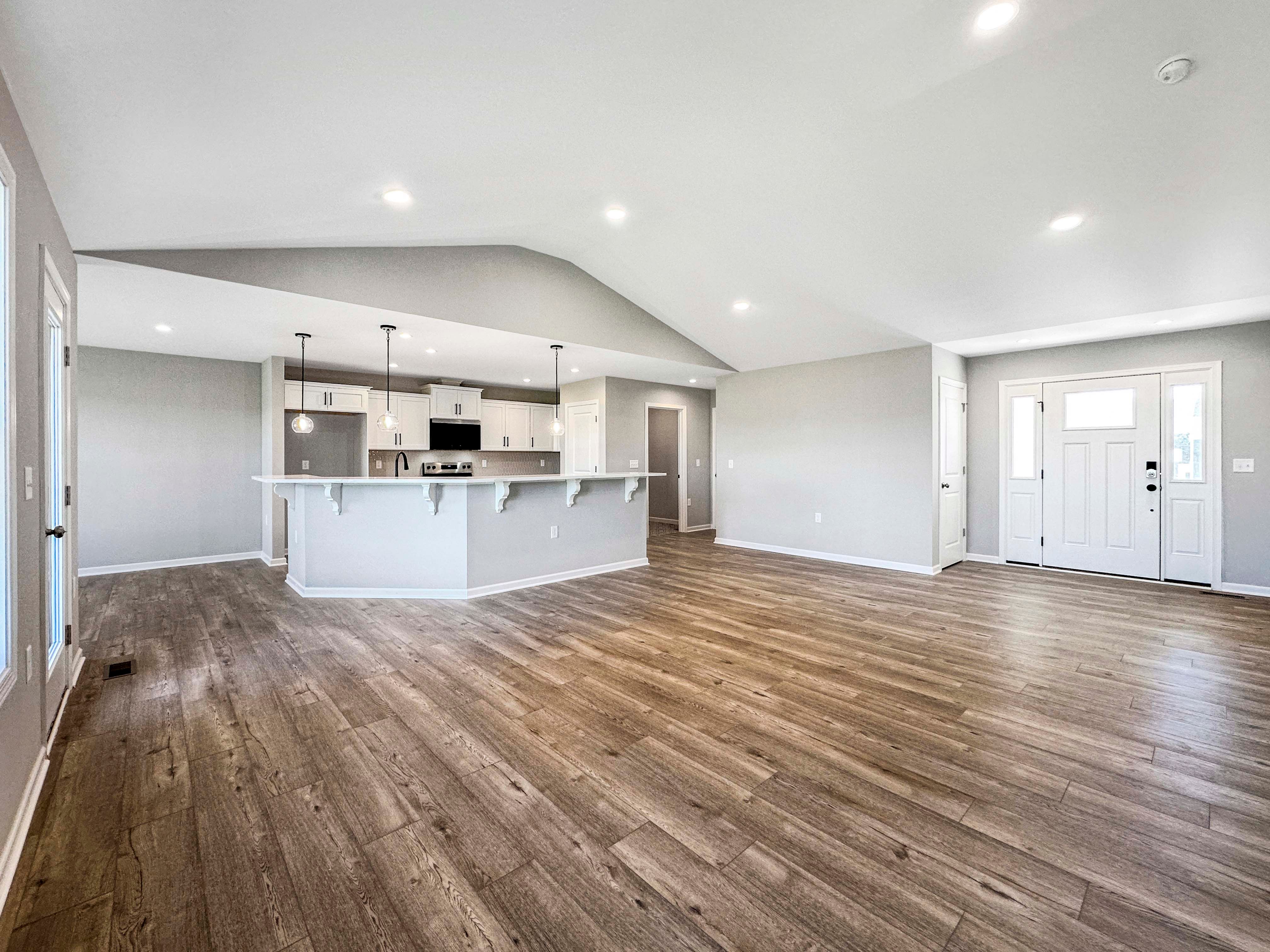 Open-concept great room and kitchen with pendant lighting, white cabinetry, and wood-like floors. Large windows provide natural light.