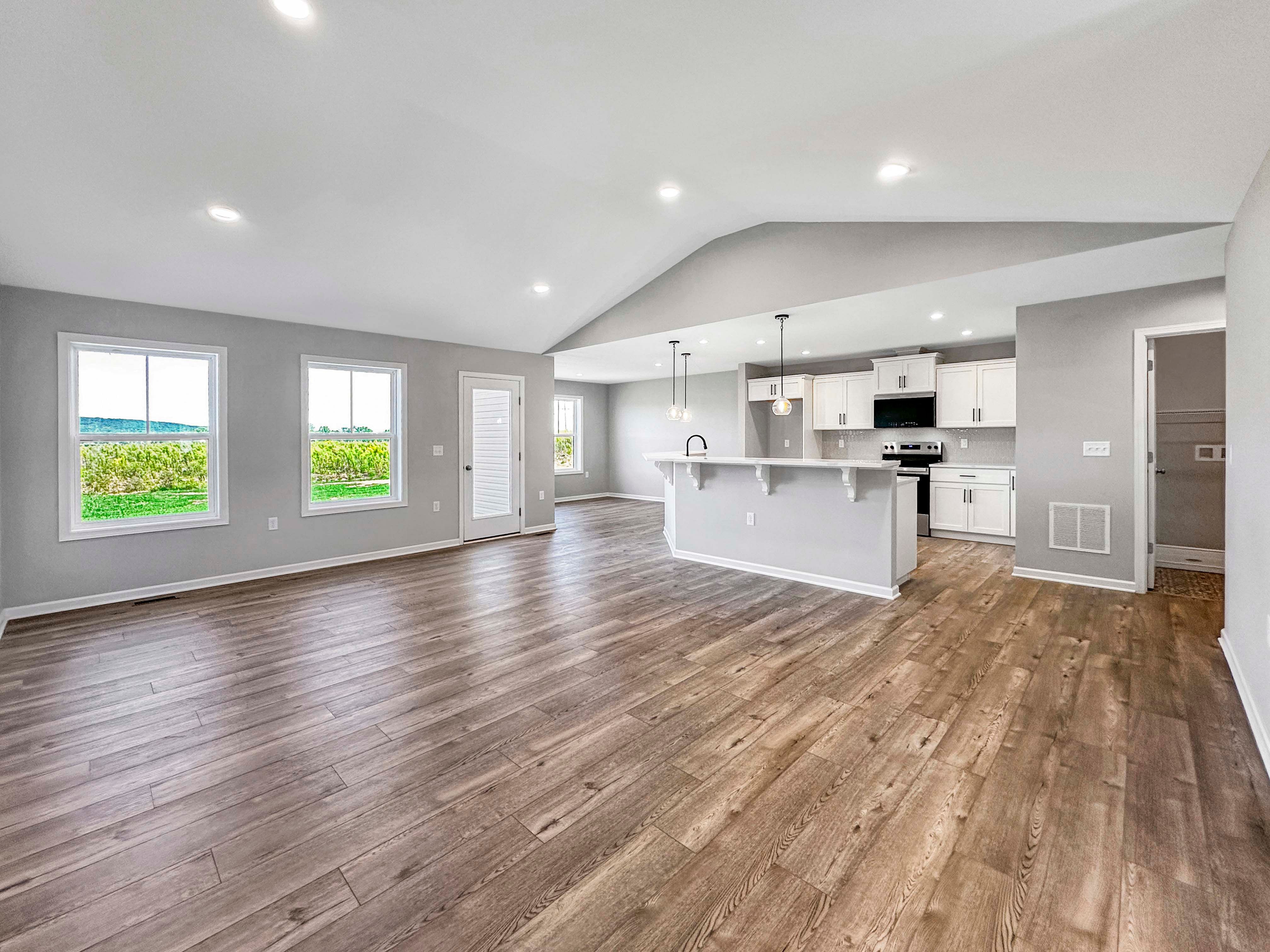 Great room with an open kitchen featuring a breakfast bar, pendant lights, and white cabinets. Large windows overlook greenery.