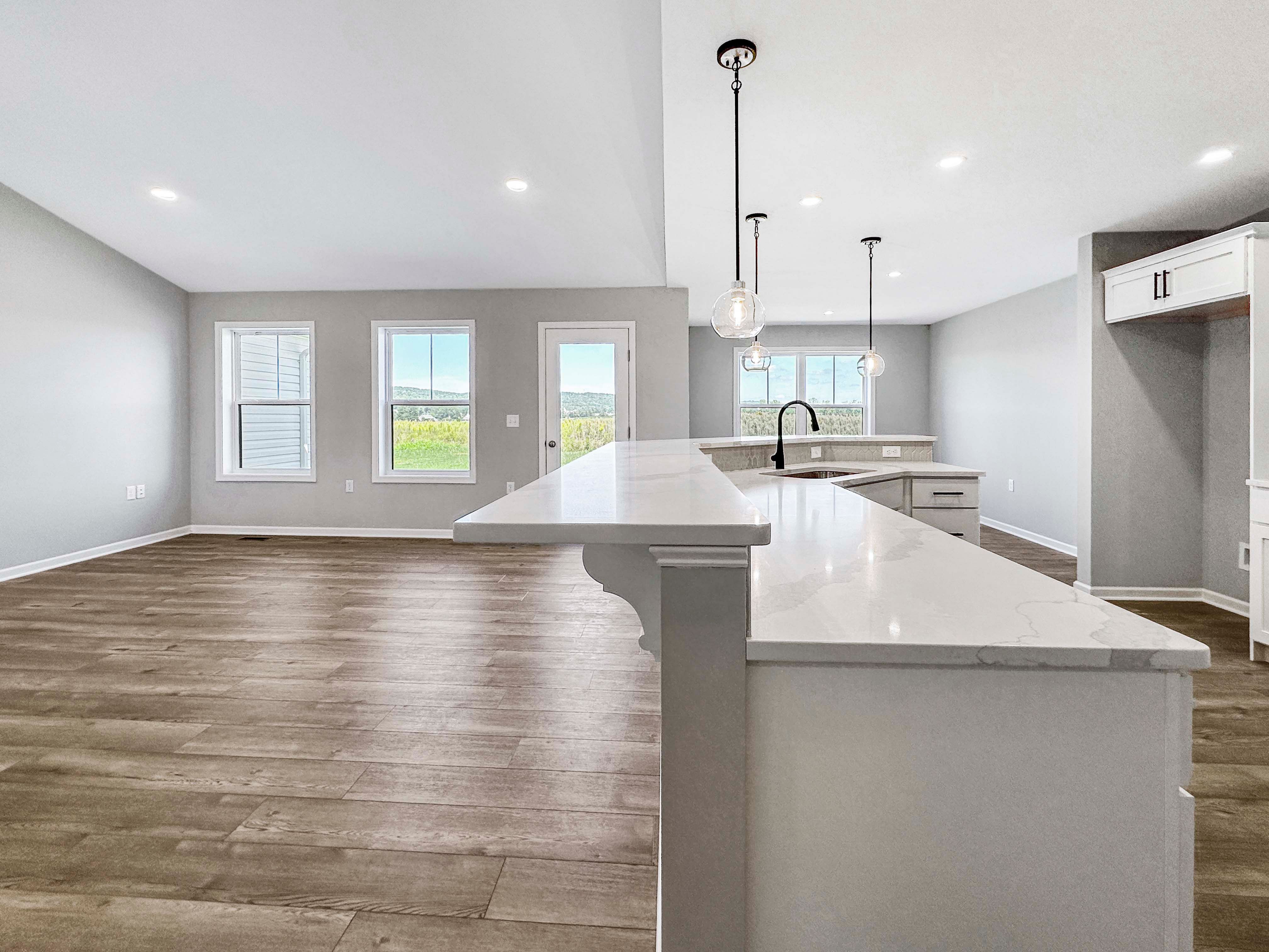 Open kitchen with white countertops, black fixtures, pendant lighting, and a view into the great room. Wood-like floors throughout.