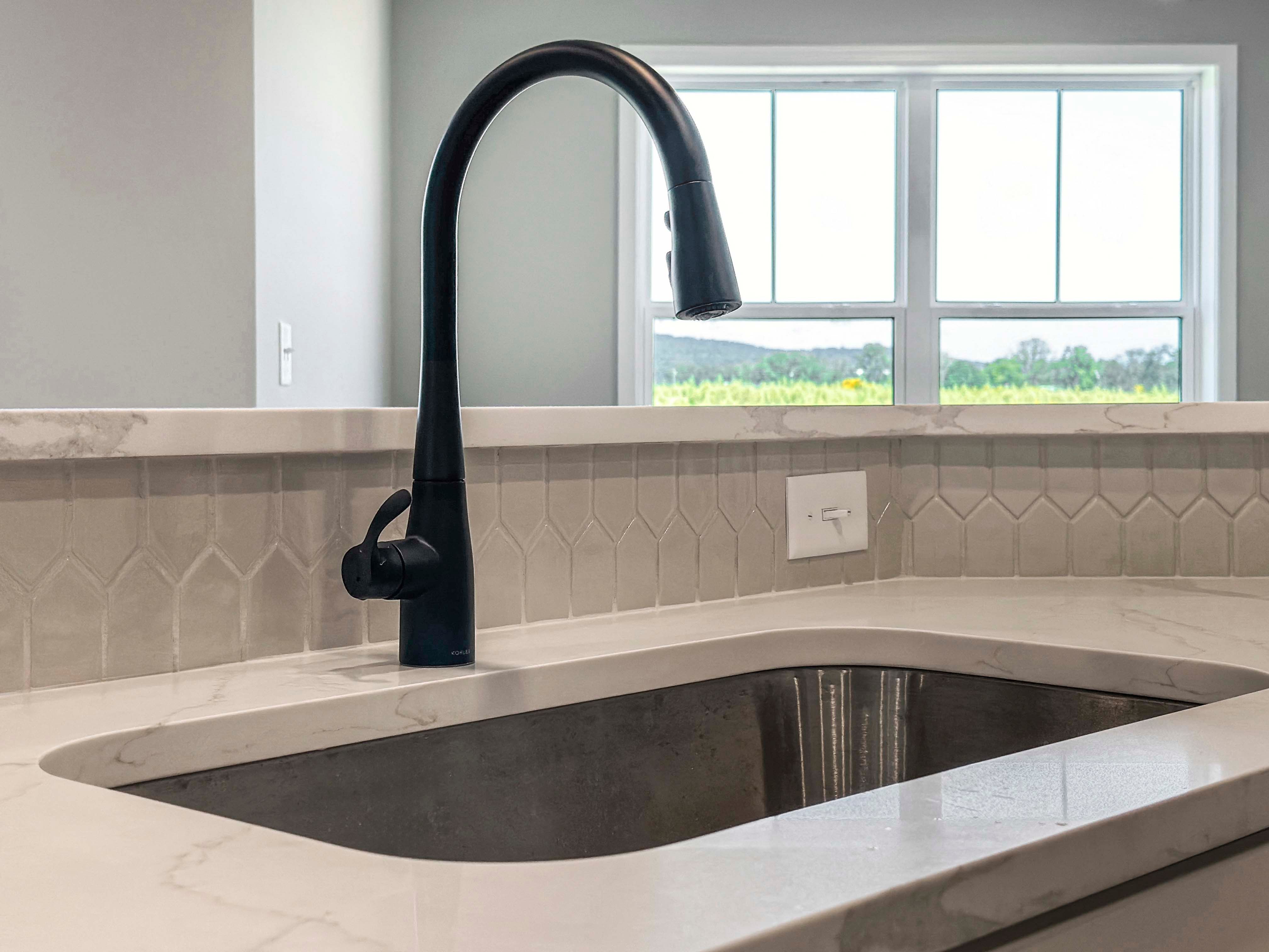 Black gooseneck faucet over a stainless steel sink, set in a marbled quartz countertop with hexagon tile backsplash and a window view.