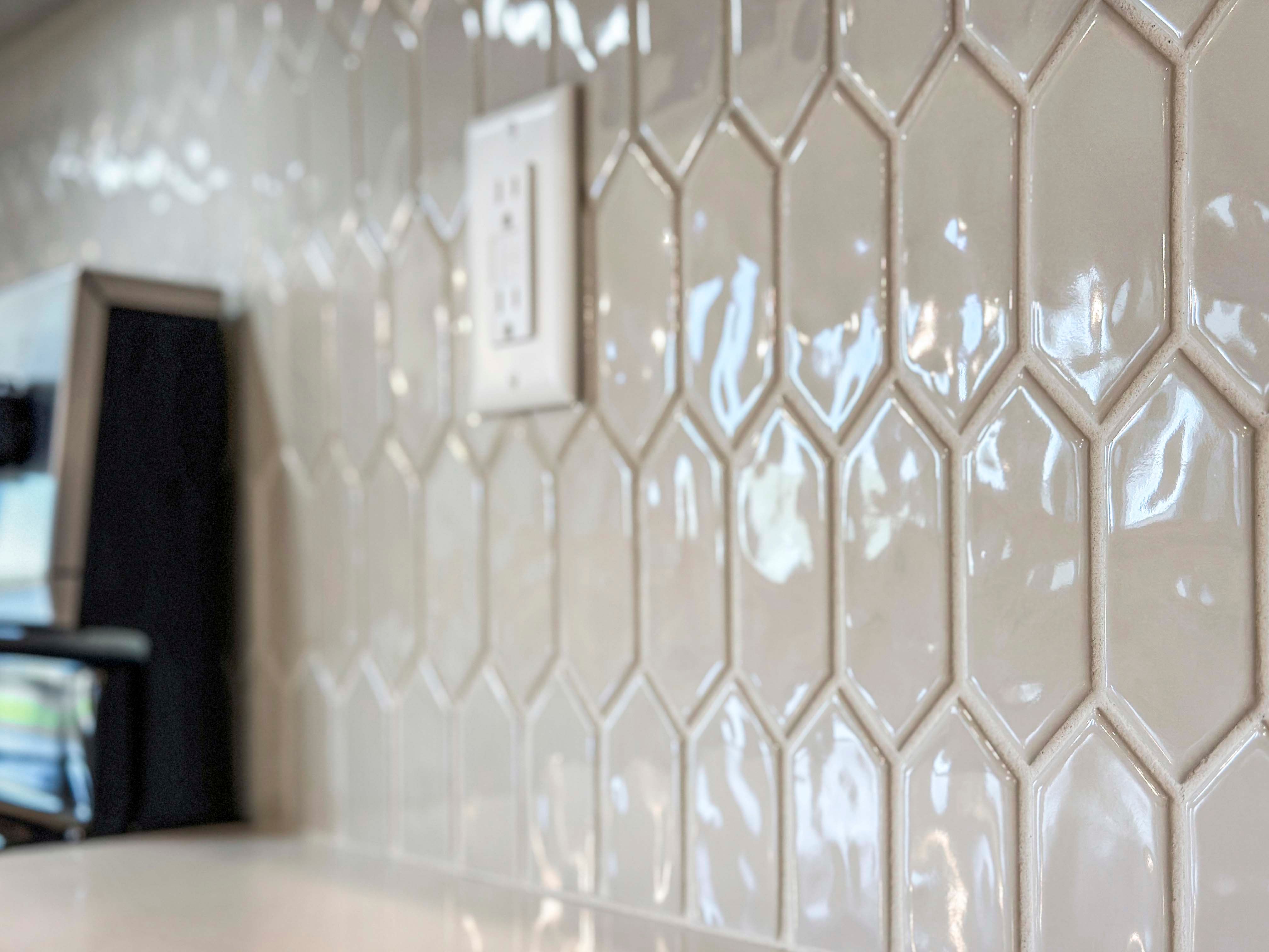 Close-up of shiny, hexagon-shaped tiles in a light gray hue, featuring an outlet integrated into the kitchen backsplash.