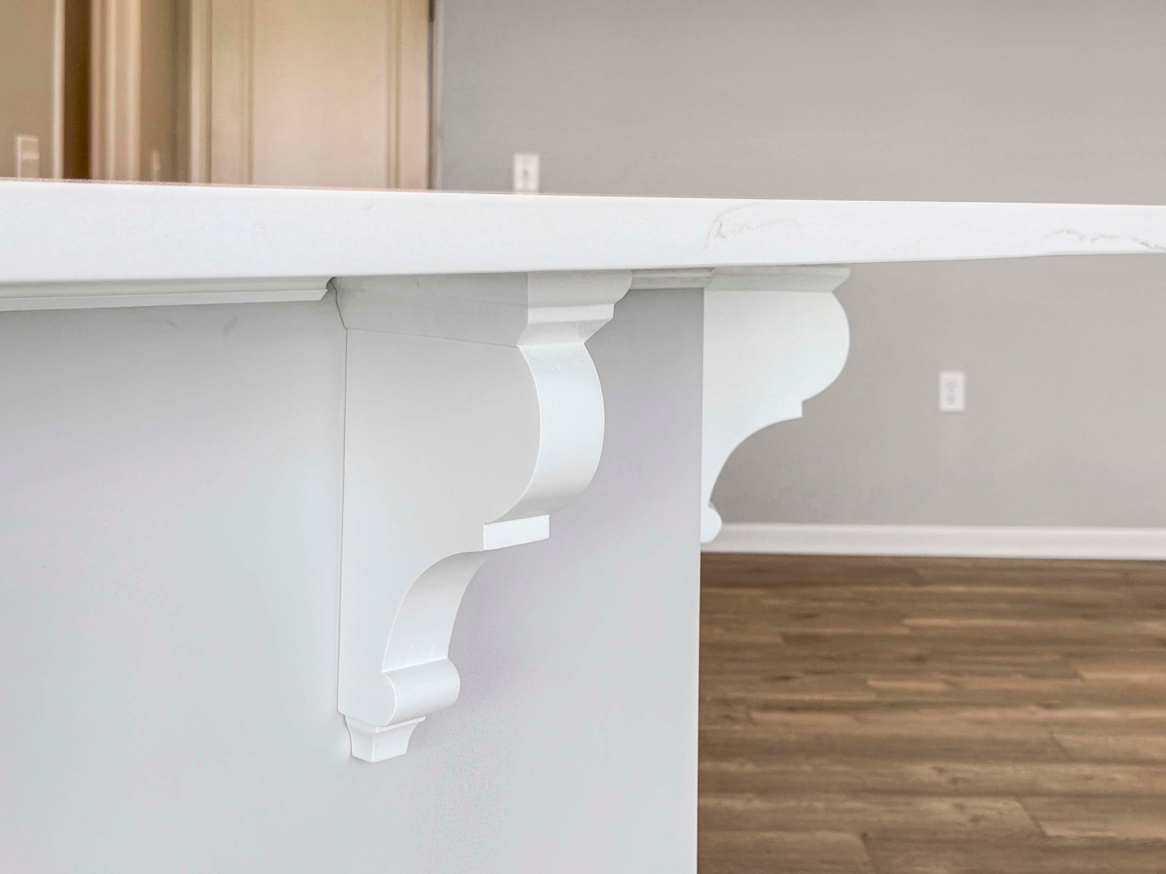 Close-up of decorative white corbels under the marbled quartz kitchen island overhang with vinyl plank flooring below.