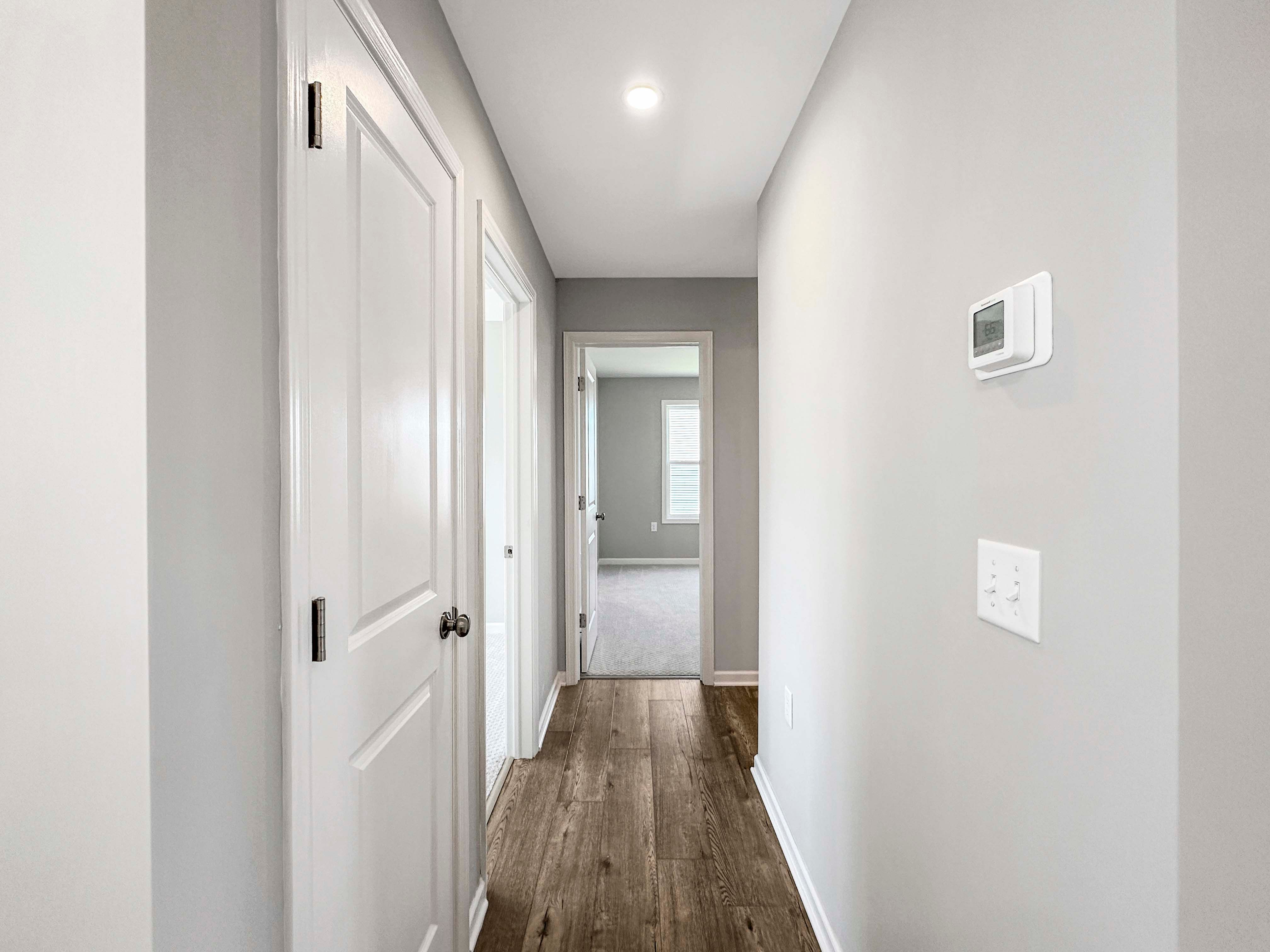 Bright hallway with wood-like flooring and white doors leading to bedrooms. Thermostat mounted on a neutral gray wall.