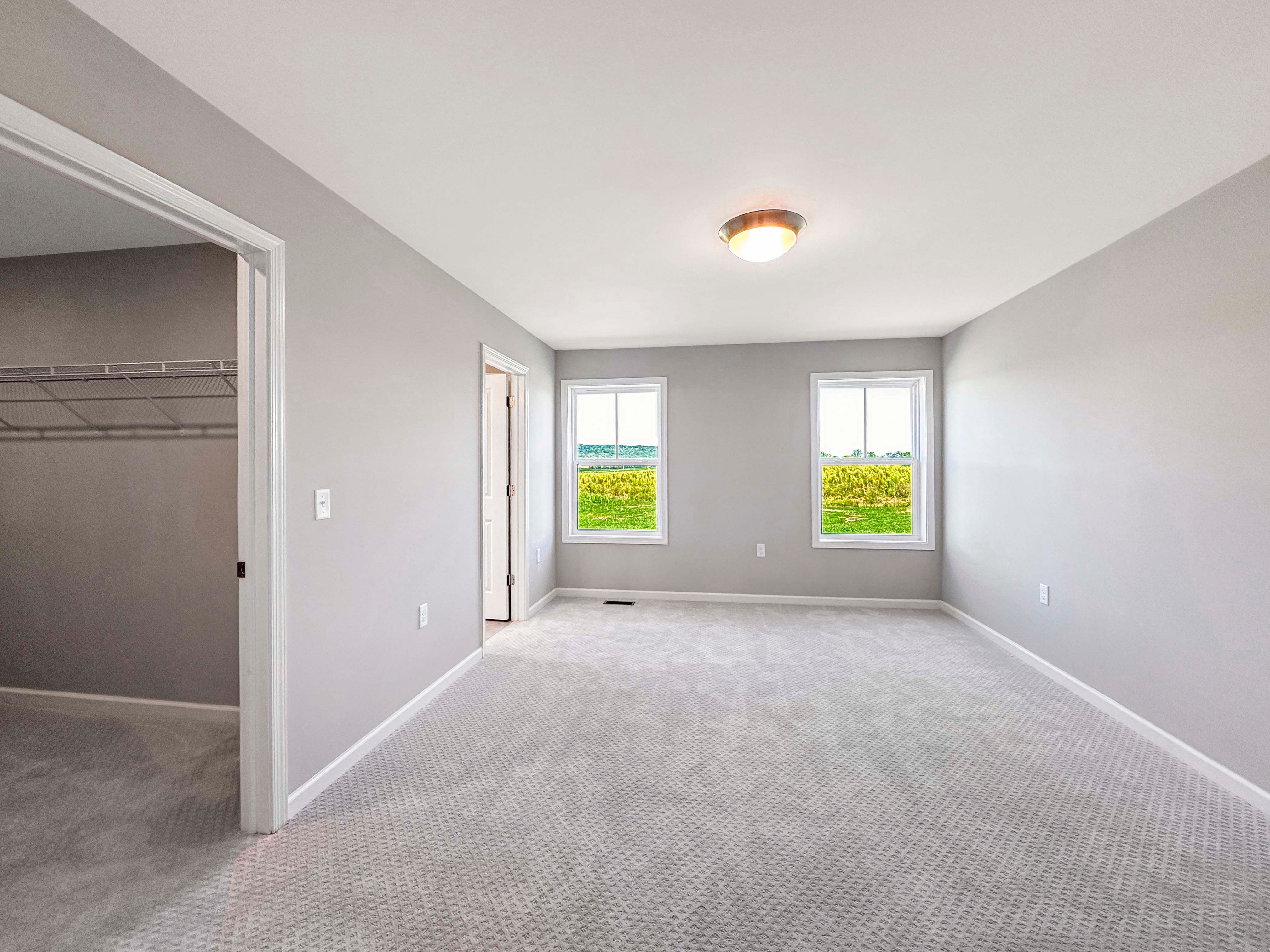 Unfurnished owners bedroom with neutral gray carpet, two windows providing natural light, and a walk-in closet entrance.
