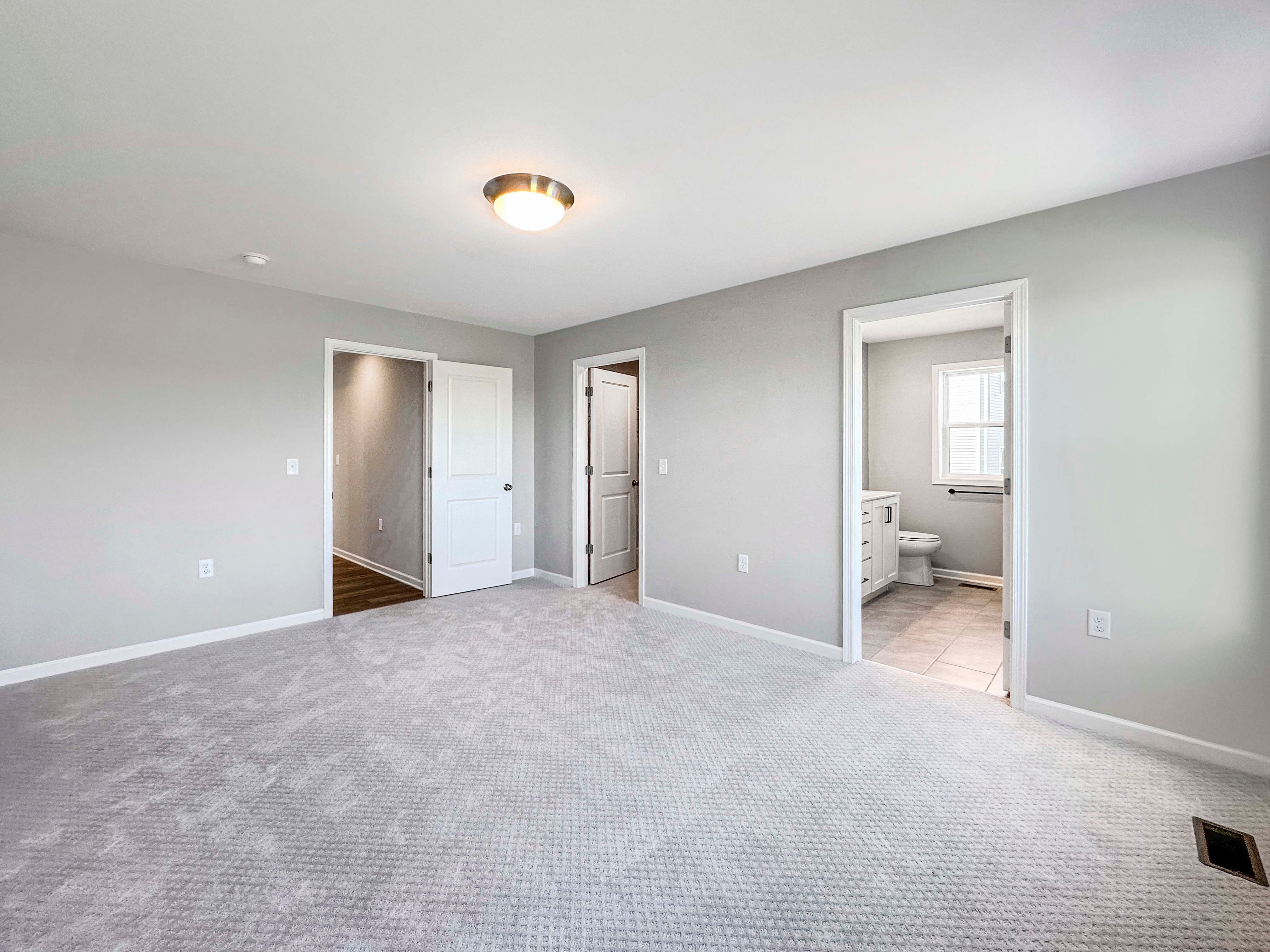 Unfurnished owners bedroom with multiple doorways, including a bathroom entrance and a walk-in closet, featuring gray carpet and soft lighting.