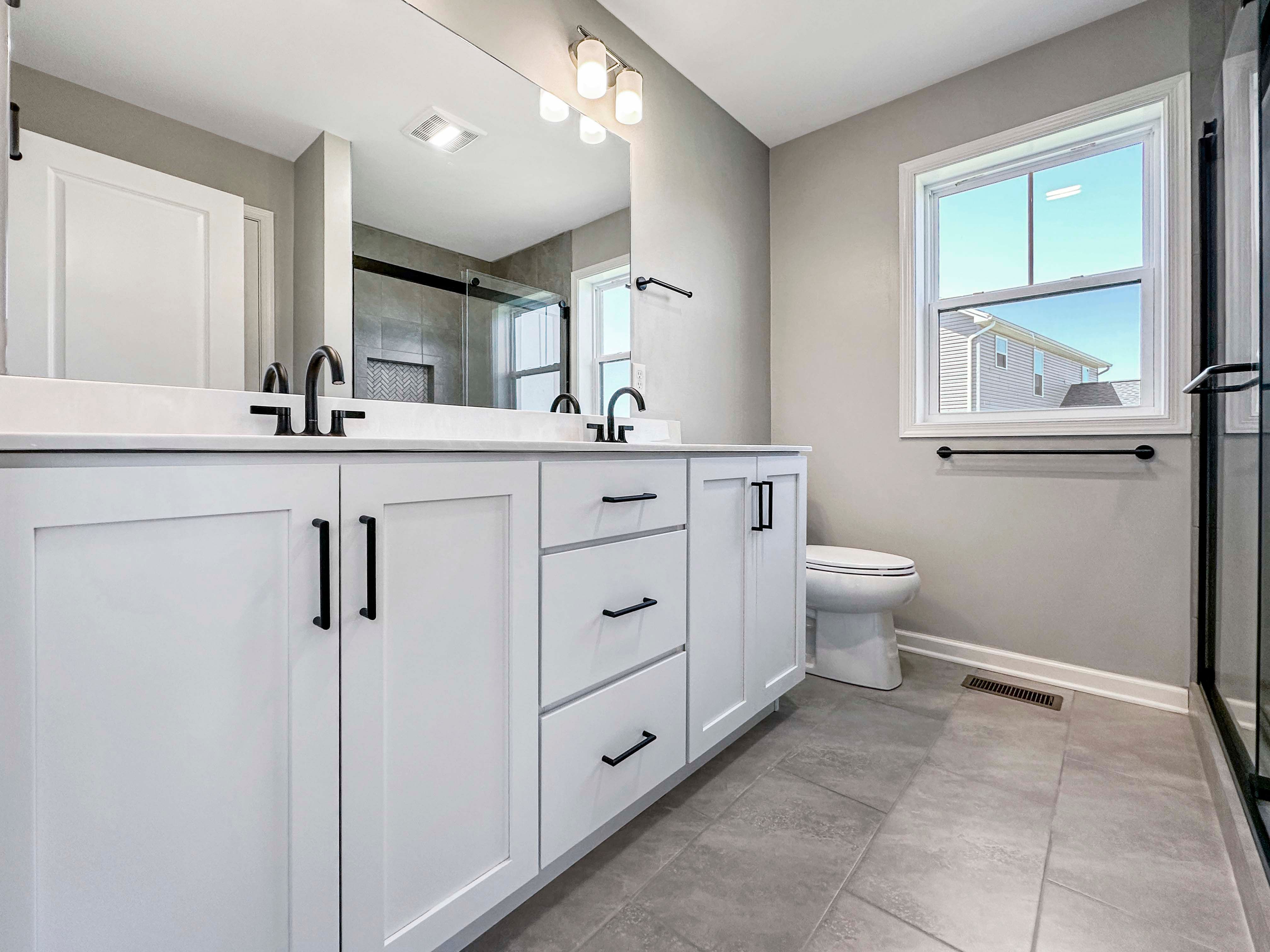 Owners bathroom showcasing a large white vanity with double sinks, black hardware, and light gray tile flooring.