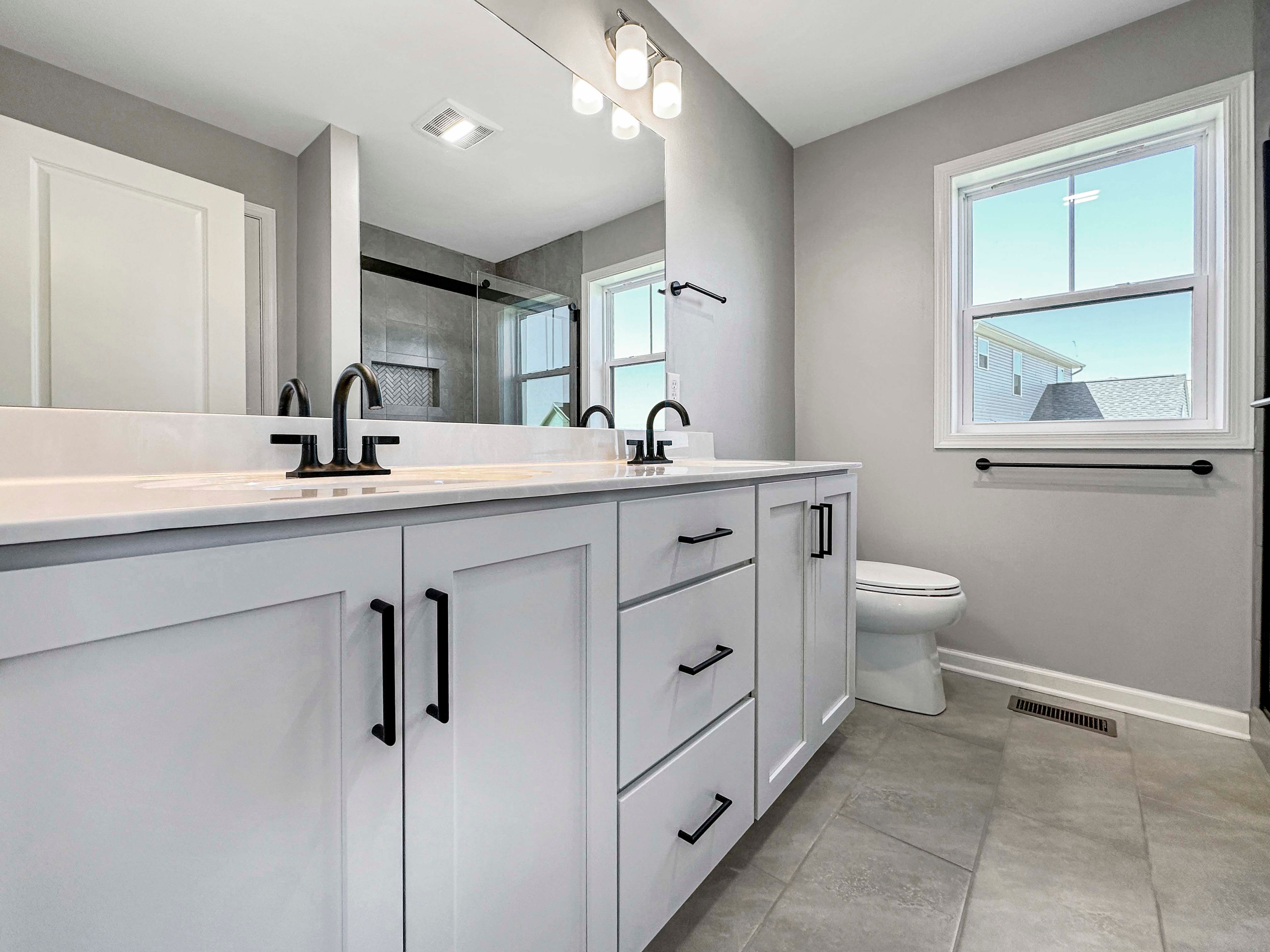Owner's bathroom with a dual-sink vanity, black faucets, large mirror, and natural light from a window.